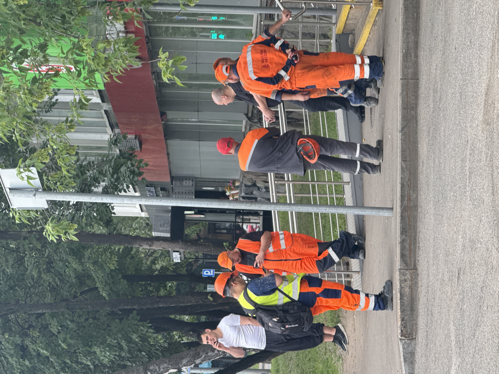 In Moscow, Russia, a group of construction workers in bright orange safety vests and pants are gathered on a sidewalk. One worker in a yellow vest and a blue shirt is looking at his phone. Another worker in an orange vest and pants is also looking at his phone. Two other workers in orange vests and pants are standing and talking. A man in a white t-shirt and black pants is standing nearby, talking on his phone. The background shows a building with a red and green facade, and trees lining the street. The scene appears to be a break or a brief stop during work.