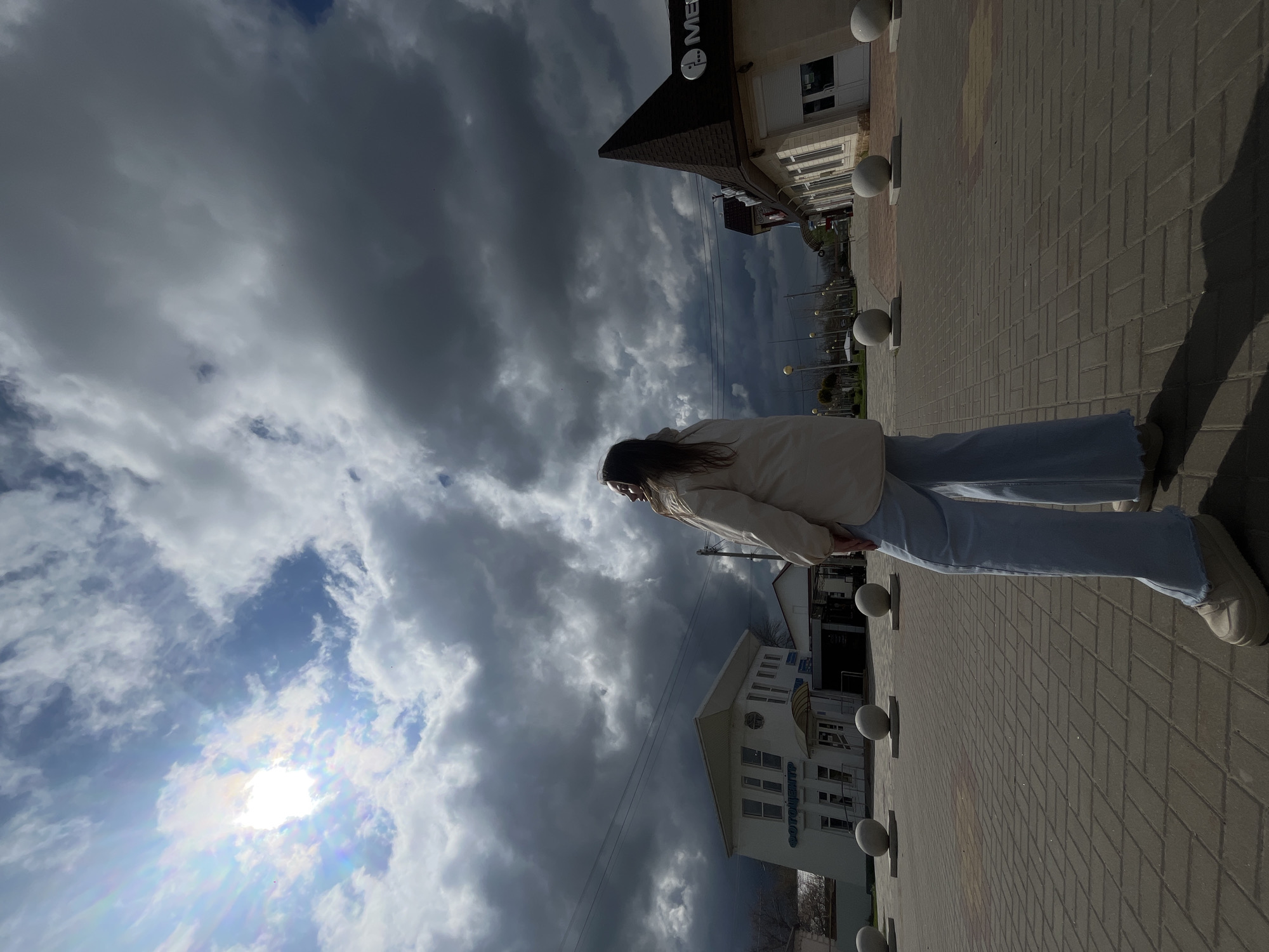 A ground-level, tilted, medium shot shows a woman standing on a pedestrian walkway in Novopokrovskaya, Russia. The walkway is made of light brown paving stones with spherical concrete bollards. The woman is facing away from the viewer. She is wearing a cream-colored jacket and light blue jeans and white sneakers. To the left and in the background are overcast skies with the sun peeking through. Buildings are also visible in the background, including one that says “Me” and another that says “ФОТОЦЕНТР”. Utility poles and power lines are visible in the background. A long shadow is cast on the walkway.