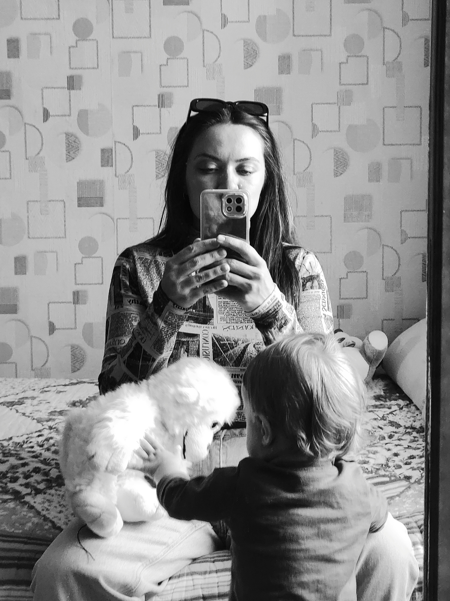 A monochrome, medium-shot photograph shows a woman and a young child reflected in a mirror. The woman holds a smartphone, presumably taking the picture, and her gaze is directed towards the phone. Sunglasses are perched on top of her head. She is wearing a patterned long-sleeved shirt. A child, facing away from the camera, reaches for a fluffy white stuffed animal which is sitting on the woman's lap. They appear to be sitting on a bed with a patterned bedspread. The wallpaper behind them is patterned with geometric shapes. The scene appears to be indoors, and based on the request, this is potentially inside a house in Chaikovsky, Russia.