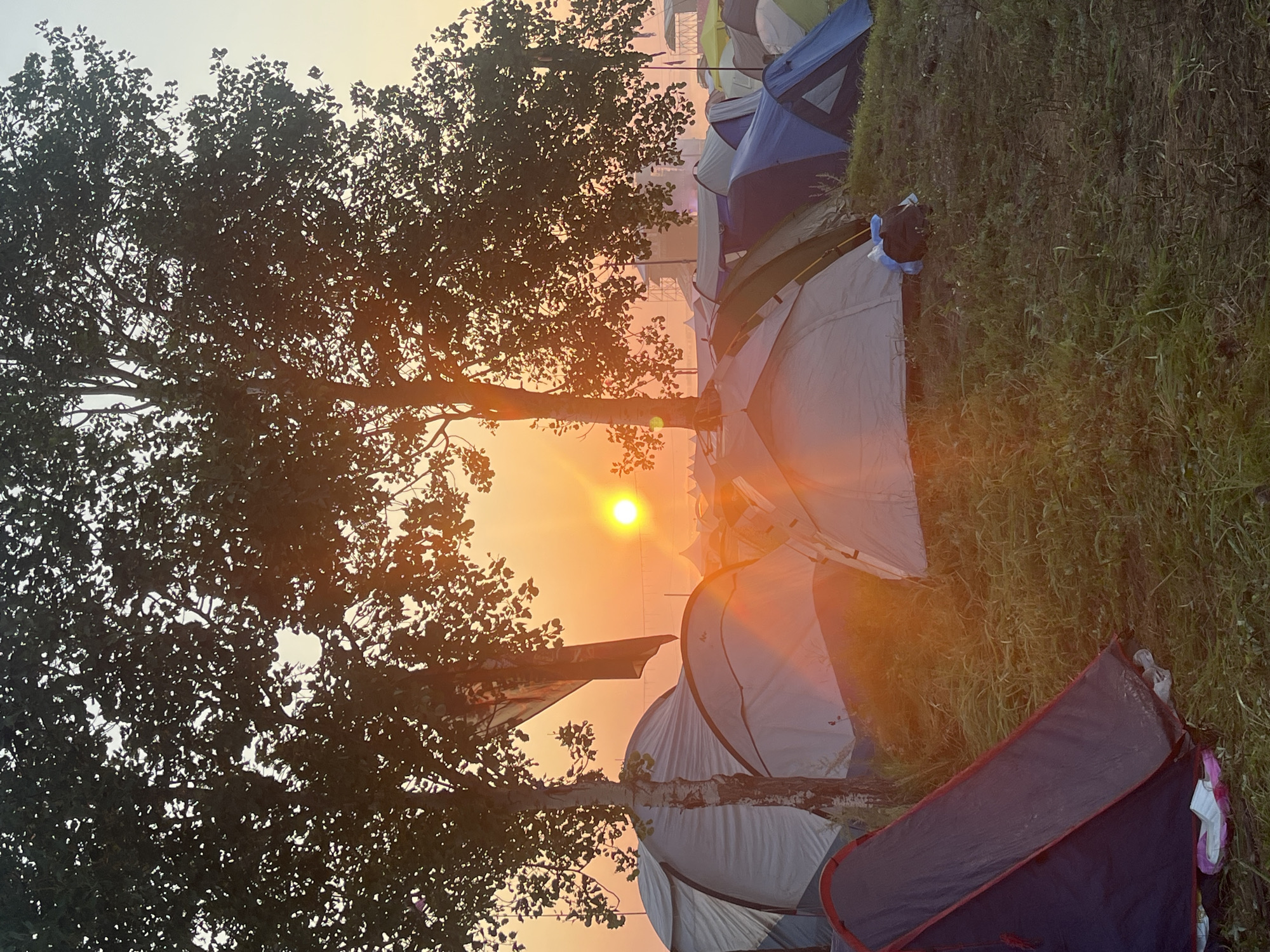The image depicts a campground at sunrise, likely in Surakovo, Russia, characterized by numerous tents pitched on grassy terrain. Two prominent trees frame the scene, their branches silhouetted against a vibrant orange and yellow sky. The sun, a bright orb, is partially obscured by the foliage of the trees, casting a warm glow and long shadows across the campsite. Tents of various colors, primarily blue, gray, and white, are visible in the foreground and middle ground. Some tents appear to be unoccupied, while others suggest recent activity with items like a blue plastic bag and white slippers visible near the tent entrances. In the distance, more tents and structures, possibly part of a larger festival or event, are faintly visible through a light haze. The overall atmosphere is peaceful and serene, capturing the quiet moments of a new day at a temporary settlement.