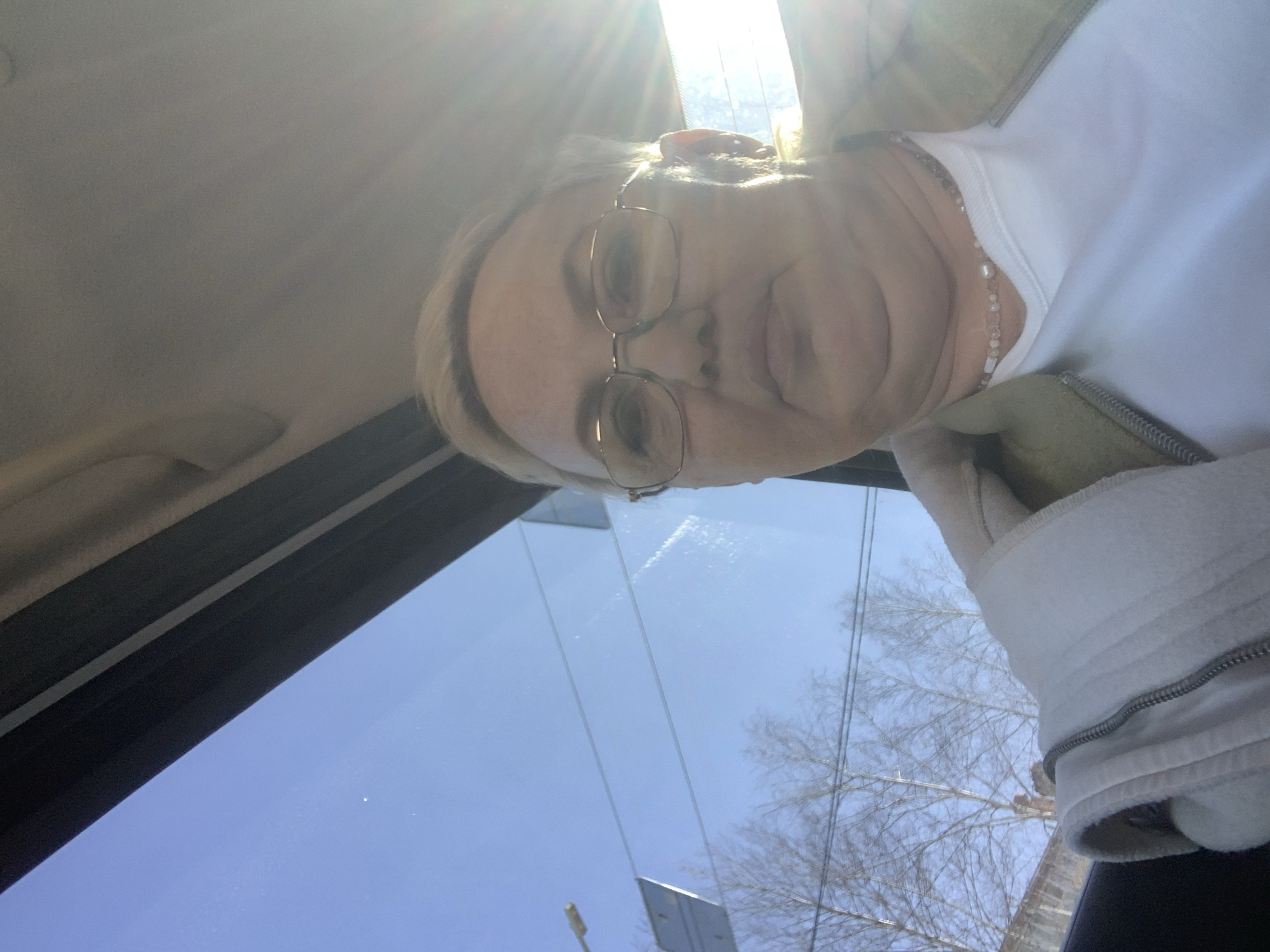 A woman with short blonde hair and glasses is looking at the camera from inside a vehicle. She is wearing a white top and a light-colored jacket with a visible zipper. Sunlight is streaming in through the windshield, and the sky is visible. Outside the window, there are bare trees and utility poles. The location is likely Chaikovsky, Russia.