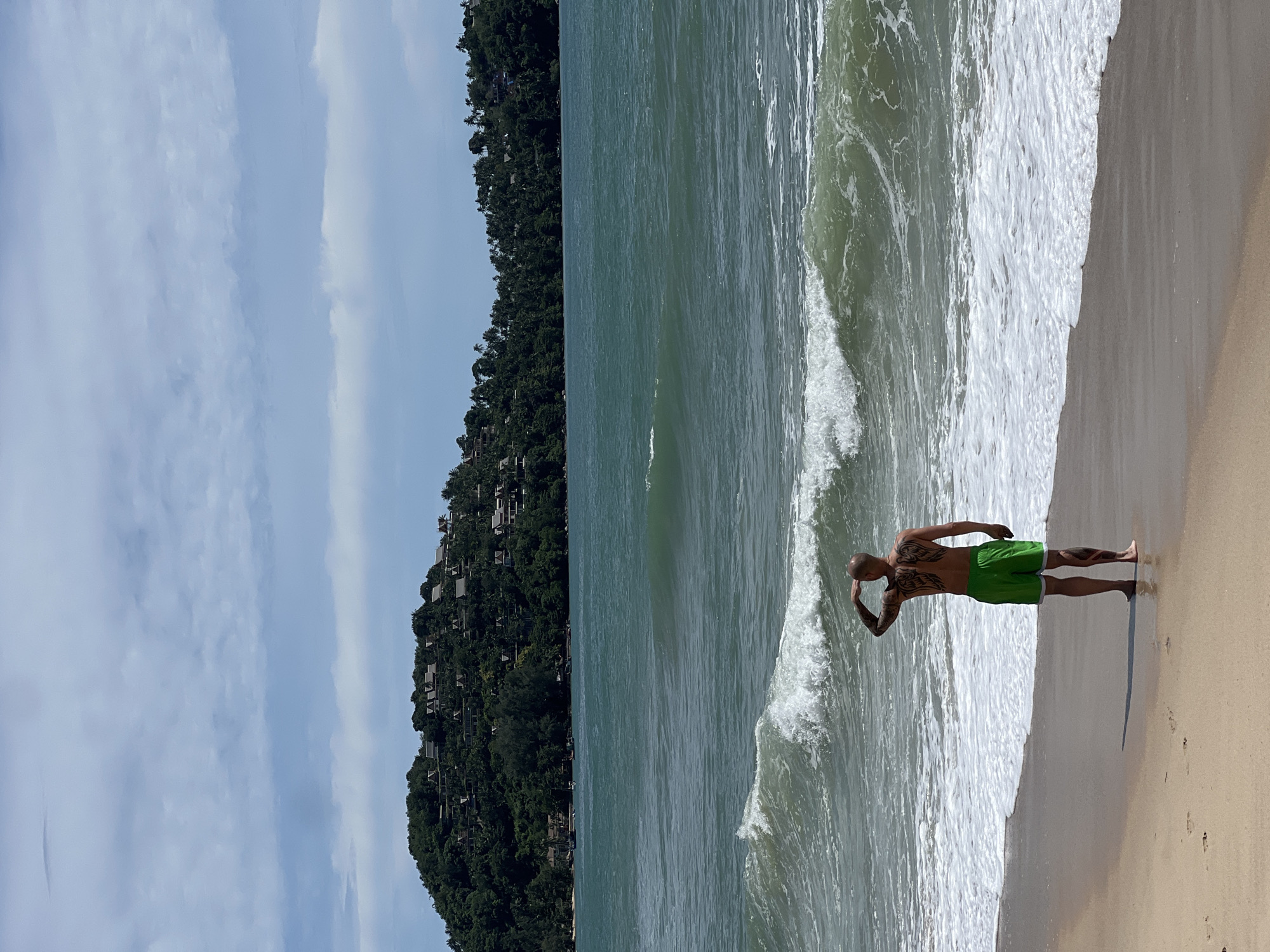 A bald, tattooed Caucasian male stands on a sandy beach, facing away from the camera and looking out at the ocean. He is wearing bright green swim shorts with white trim and is barefoot on the wet sand where waves recede. His right hand is raised to his forehead, shielding his eyes as he gazes towards the horizon. Visible on his back are large, dark tattoos resembling wings, and further tattoos are present on his lower legs. A distinct shadow is cast behind him on the sand.
The ocean in the midground features multiple breaking waves with white foam, exhibiting a turquoise to deep blue-green hue. In the background, a large, lush green hillside, densely covered with trees and tropical vegetation, rises across the water. Numerous buildings with light-colored roofs, possibly villas or resort structures, are interspersed within the foliage on the hillside. The sky above is light blue with scattered stratocumulus clouds. The scene is brightly lit under what appears to be a sunny day in an unknown coastal location.