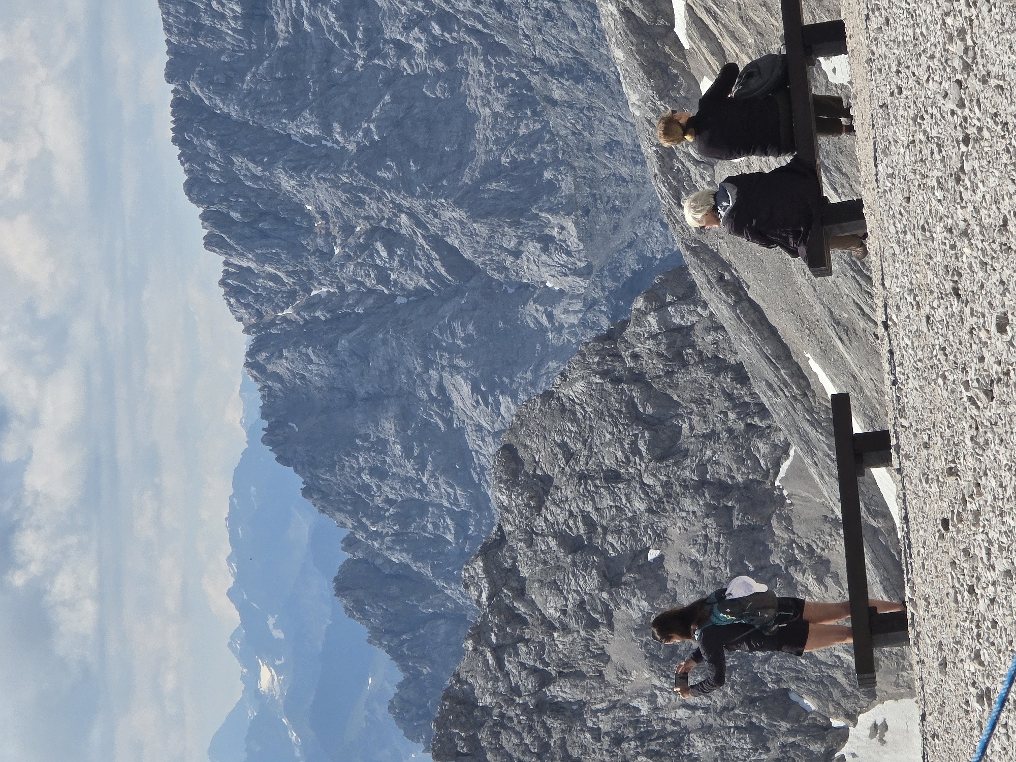The image depicts a mountainous landscape with three people. In the foreground to the left, a woman with long dark hair is standing and holding a smartphone up to take a picture. She is wearing a dark jacket, black shorts, and a backpack. In the mid-ground to the right, two individuals are seated on a dark bench. The person on the left has short, light-colored hair and is wearing a dark jacket. The person on the right has light brown hair tied back and is wearing a dark jacket. The background features rugged, rocky mountains under a partly cloudy sky. The terrain in the foreground is covered in small stones and gravel. The scene is likely from the Zugspitze, the highest mountain in Germany, which is near Garmisch-Partenkirchen.