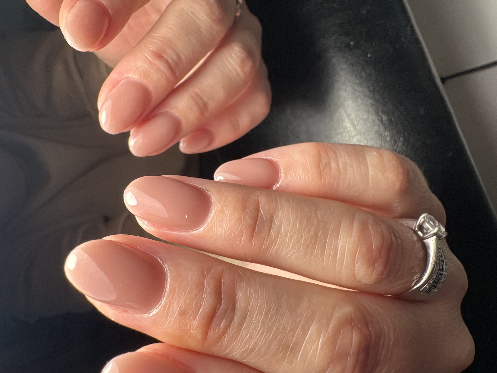 A close-up image shows two hands with freshly manicured nails, set against a dark, indistinct background. The hand in the foreground is sharply focused, displaying nails painted in a glossy, light nude or pale pink shade with a soft oval or almond shape. The skin on the fingers exhibits natural lines and texture. A silver-toned ring adorned with clear stones and a textured band, possibly a Greek key pattern, is visible on the ring finger of this hand.
The second hand is positioned in the upper background, partially visible and slightly out of focus, also featuring similar manicured nails. The lighting accentuates the shine on the nails and the subtle contours of the hands. The scene is likely an indoor setting, possibly after a beauty treatment. No specific activity is taking place other than the presentation of the nails. There is no visible text. The overall appearance suggests personal grooming and attention to detail. This scene is observed in Chaikovsky, Russia.