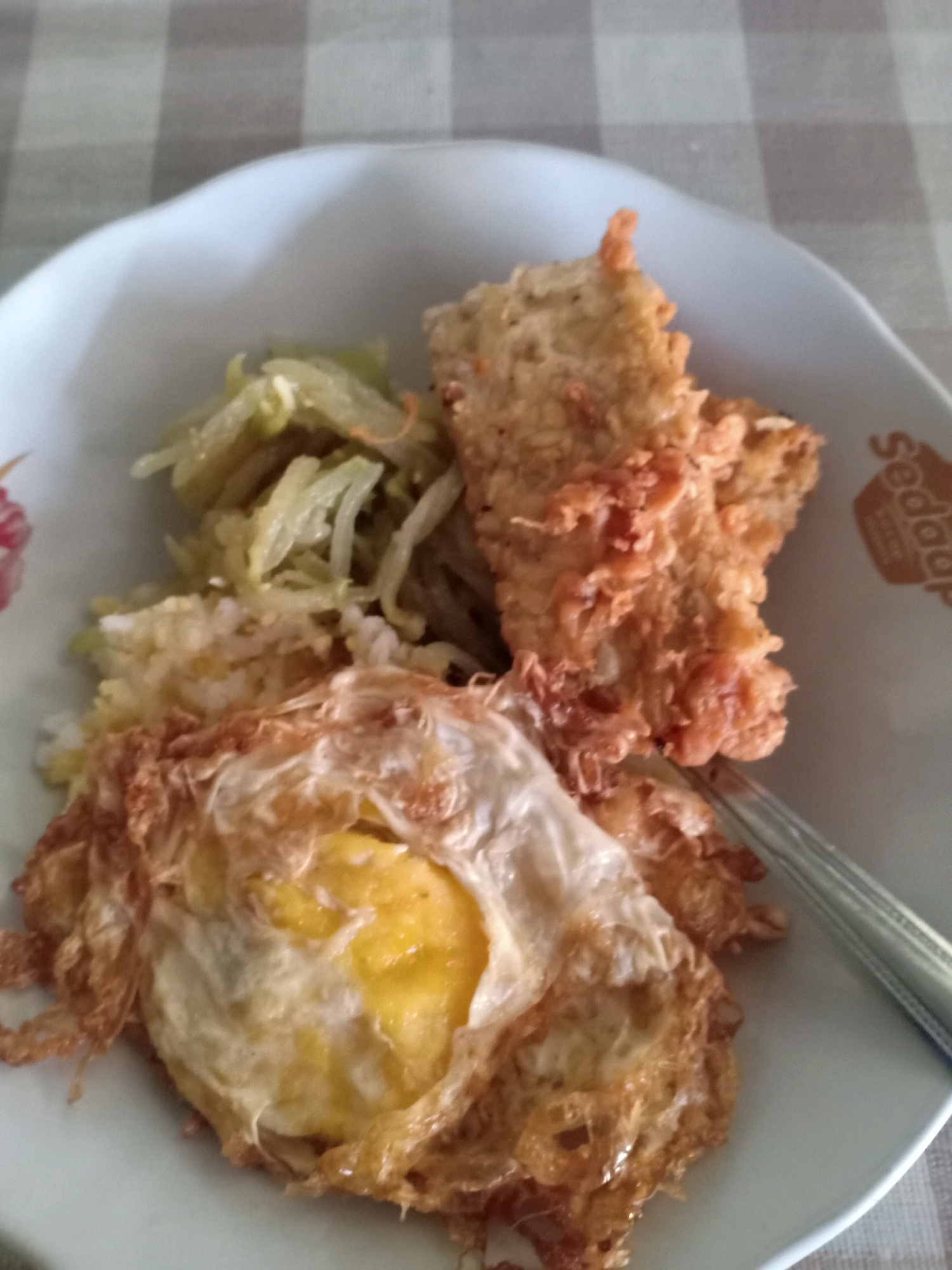 An overhead close-up shot shows a white bowl of food placed on a checkered tablecloth. The bowl contains rice, a fried egg, a portion of fried chicken, and shredded vegetables. The fried egg is cooked sunny-side up with a visible yellow yolk. The fried chicken appears crispy with a golden-brown coating. The shredded vegetables are light green and appear to be seasoned. A fork is visible on the right side of the bowl, partially submerged in the food. The background consists of the checkered tablecloth and the edge of the white bowl. The image appears to be taken in a home setting in Sumberrejo, Indonesia, possibly a dining table during a meal.
