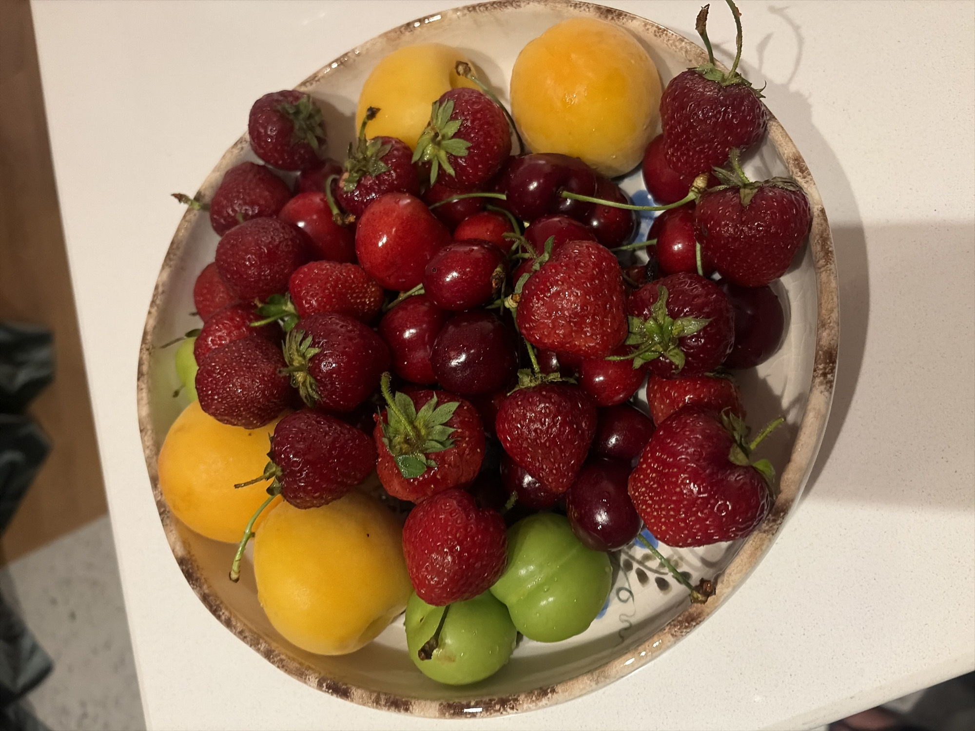 A shallow, decorative bowl filled with assorted fruits sits on a white, speckled countertop. The bowl has a brown rim with a textured pattern and a white interior adorned with blue floral motifs. The fruits include bright red strawberries, dark red cherries, green plums, and yellow plums or apricots. The arrangement is densely packed, with the fruits spilling over the edges of the bowl. The lighting suggests an indoor setting, possibly a kitchen, with a shadow cast on the countertop from the bowl. The background is blurred, showing a hint of dark furniture or cabinetry. There are no people or actions depicted in the image. The scene appears to be a still life of fresh produce.