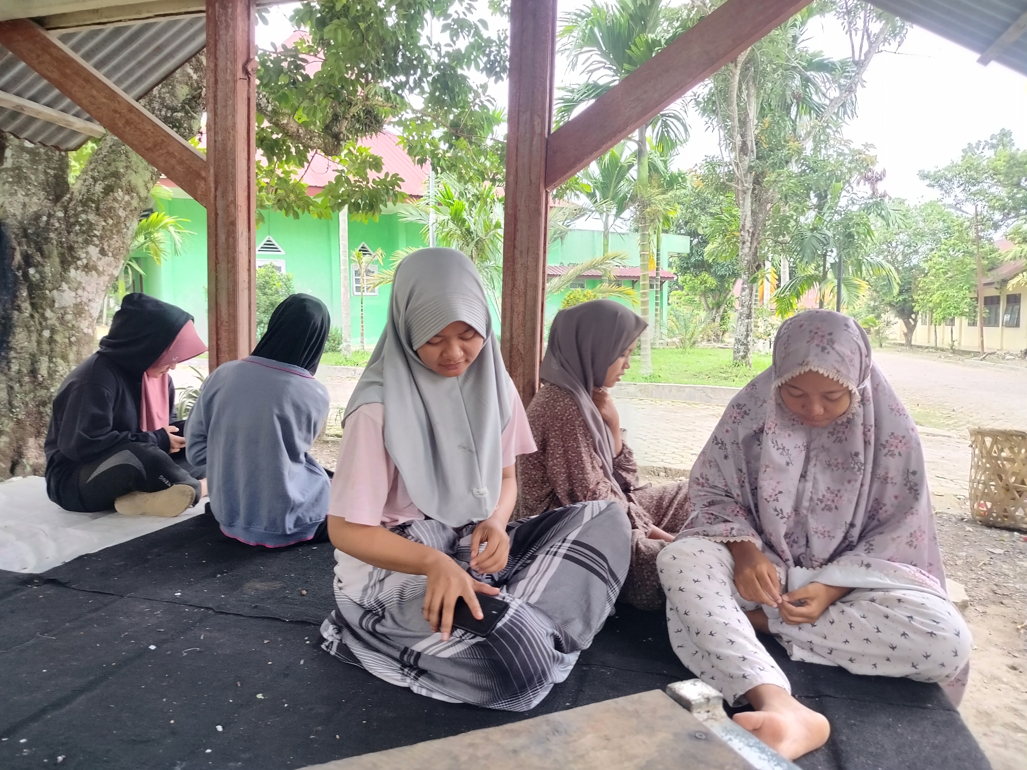 Four individuals, all wearing hijabs and traditional Islamic attire, are seated on a black mat outdoors under a covered structure. The structure has a corrugated metal roof supported by wooden beams and a reddish-brown metal column. In the background, a bright green building with white-framed windows is visible, along with trees and other foliage. The ground is a mixture of dirt and paved areas. The individuals are positioned from left to right, with the first two in darker clothing, one in a black hoodie and pink hijab, and the other in a grey top and black hijab. The third individual is wearing a light grey hijab and a patterned sarong, and is holding a black smartphone. The fourth individual on the far right is wearing a patterned hijab and white pants with a bird motif, and appears to be looking down at her hands. To the right of the fourth individual, a woven basket is visible. The scene suggests a casual gathering or resting spot, potentially in a school or community area, given the presence of the buildings and open space. The city of Purwodadi, Indonesia, is the geographical context for this image.