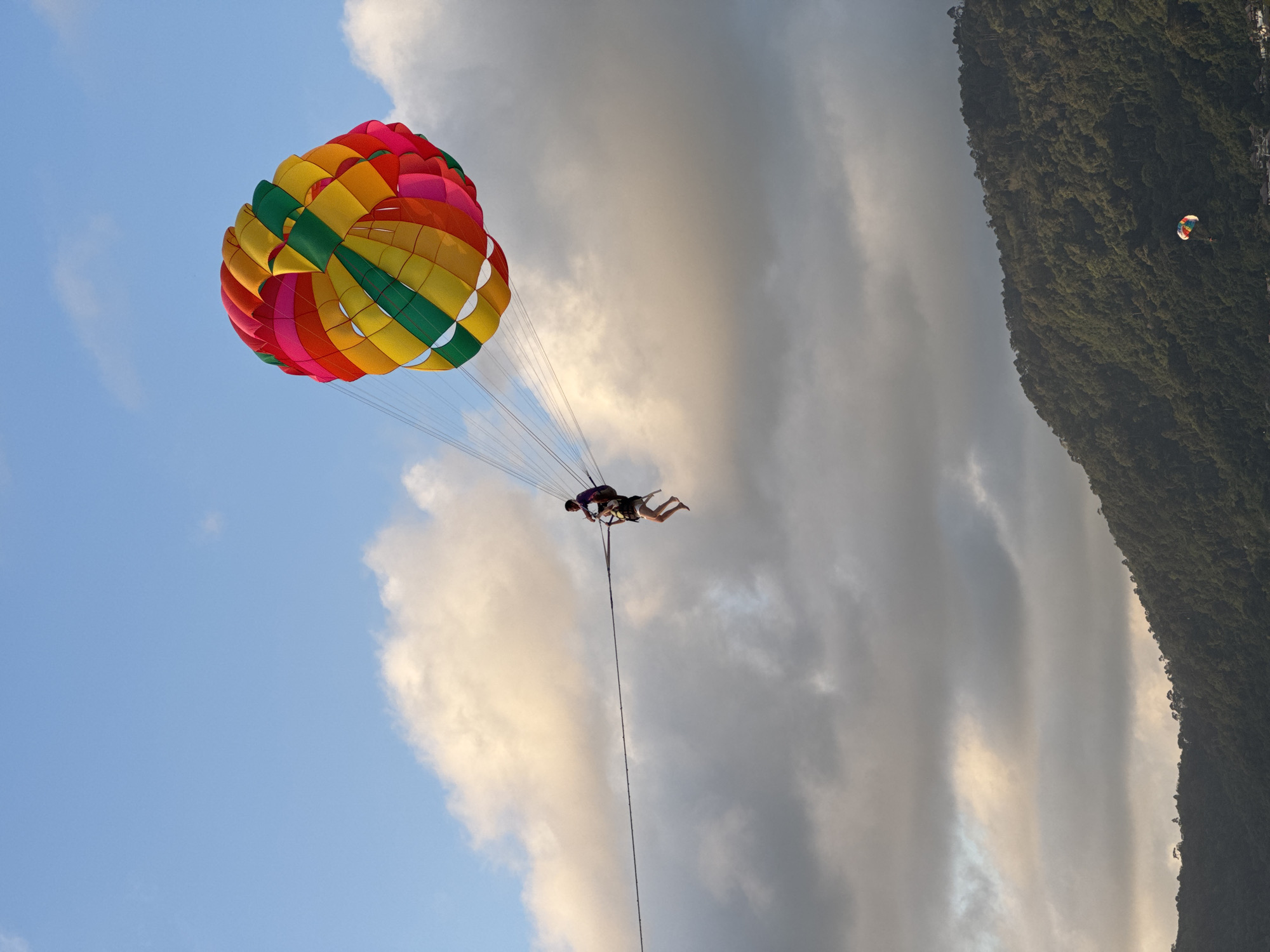 In Patong, Thailand, two individuals are parasailing against a backdrop of a cloudy sky and a tree-covered mountainside. The parasail is a vibrant sphere of red, pink, yellow, and green, suspended by thin lines. Below the parasail, two people are seated in a harness, one appears to be steering while the other is seated behind. They are attached to a tow rope that stretches horizontally across the frame. The mountain is lush with green trees, and a hint of buildings can be seen at its base. The sun is setting, casting a warm glow on the clouds and illuminating the scene.