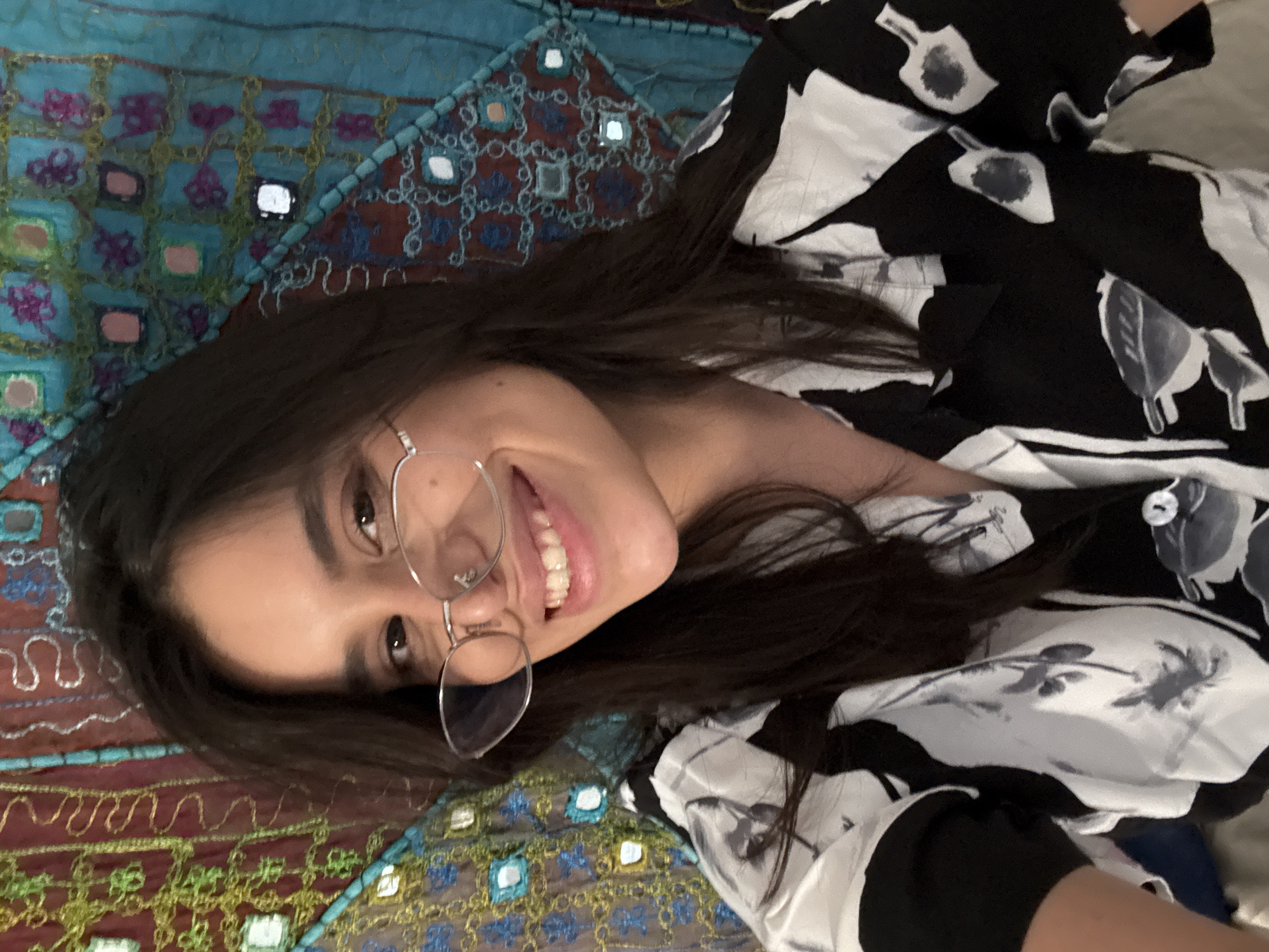 A young woman with dark hair and glasses smiles at the camera. She is wearing a black and white patterned shirt. The background is a colorful tapestry with intricate patterns and small mirror embellishments. The image is a close-up selfie. There is no identifiable location in the city of Las Rozas de Madrid, Spain.