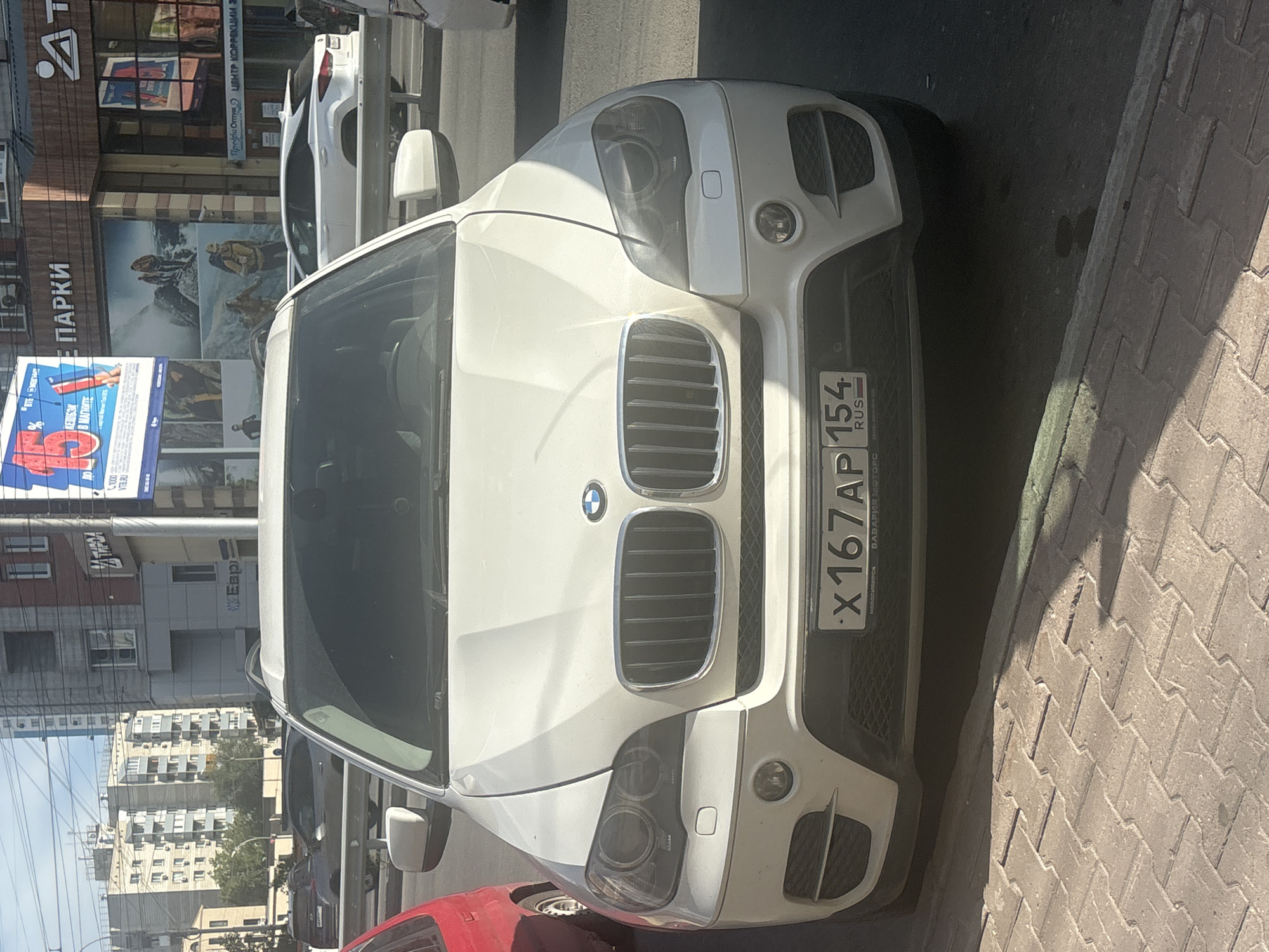A white BMW X5 is parked on a street in Novosibirsk, Russia, on a sunny day. The car's license plate reads "X167AP 154 RUS". The car is facing the camera and is parked on a sidewalk with patterned paving stones. In the background, there are buildings, including an apartment building and a commercial building with a large advertisement for an outdoor gear store. Other vehicles are visible on the street behind the BMW. A red car is parked to the left of the BMW. The sky is clear and blue.