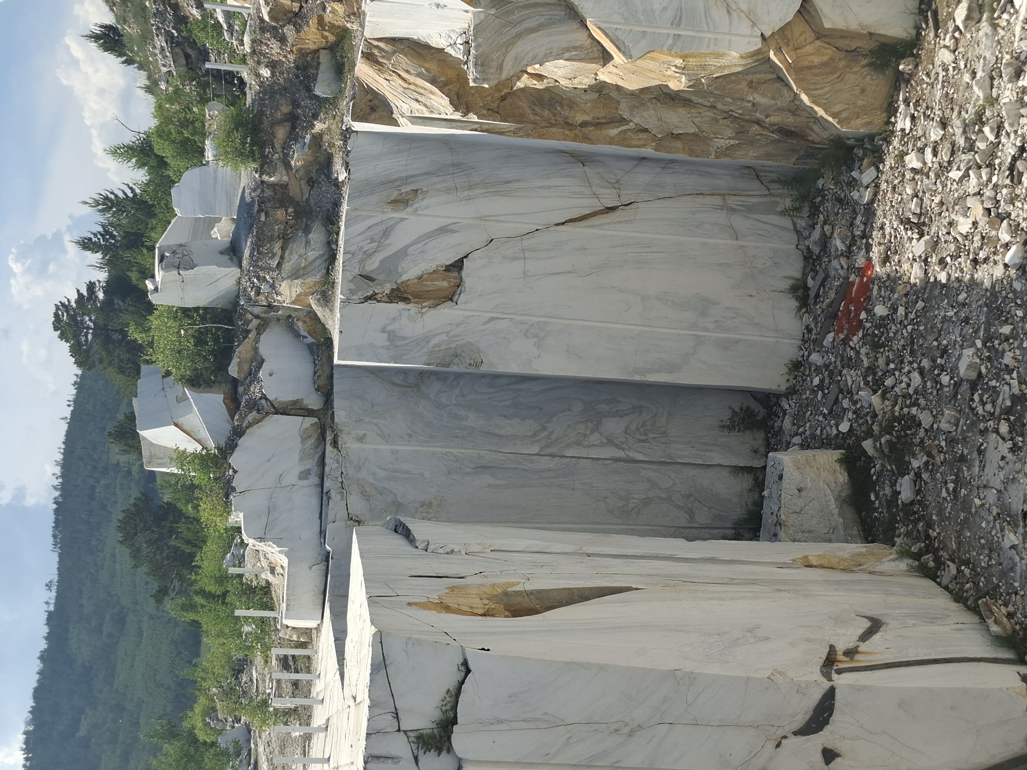 This media file depicts a marble quarry in Bugul'deika, Russia. The scene is dominated by massive, stacked blocks of white marble, some of which are fractured, revealing veins of tan and gray. The marble is cut into rough, geometric shapes, suggesting it's in the process of extraction or preparation. In the background, a lush, green hillside covered in trees rises steeply under a bright, partly cloudy sky. The overall impression is one of raw nature and industrial activity, with the quarry starkly contrasting with the verdant landscape. The time of day appears to be daytime, with clear sunlight casting shadows. There is no visible text in the image.