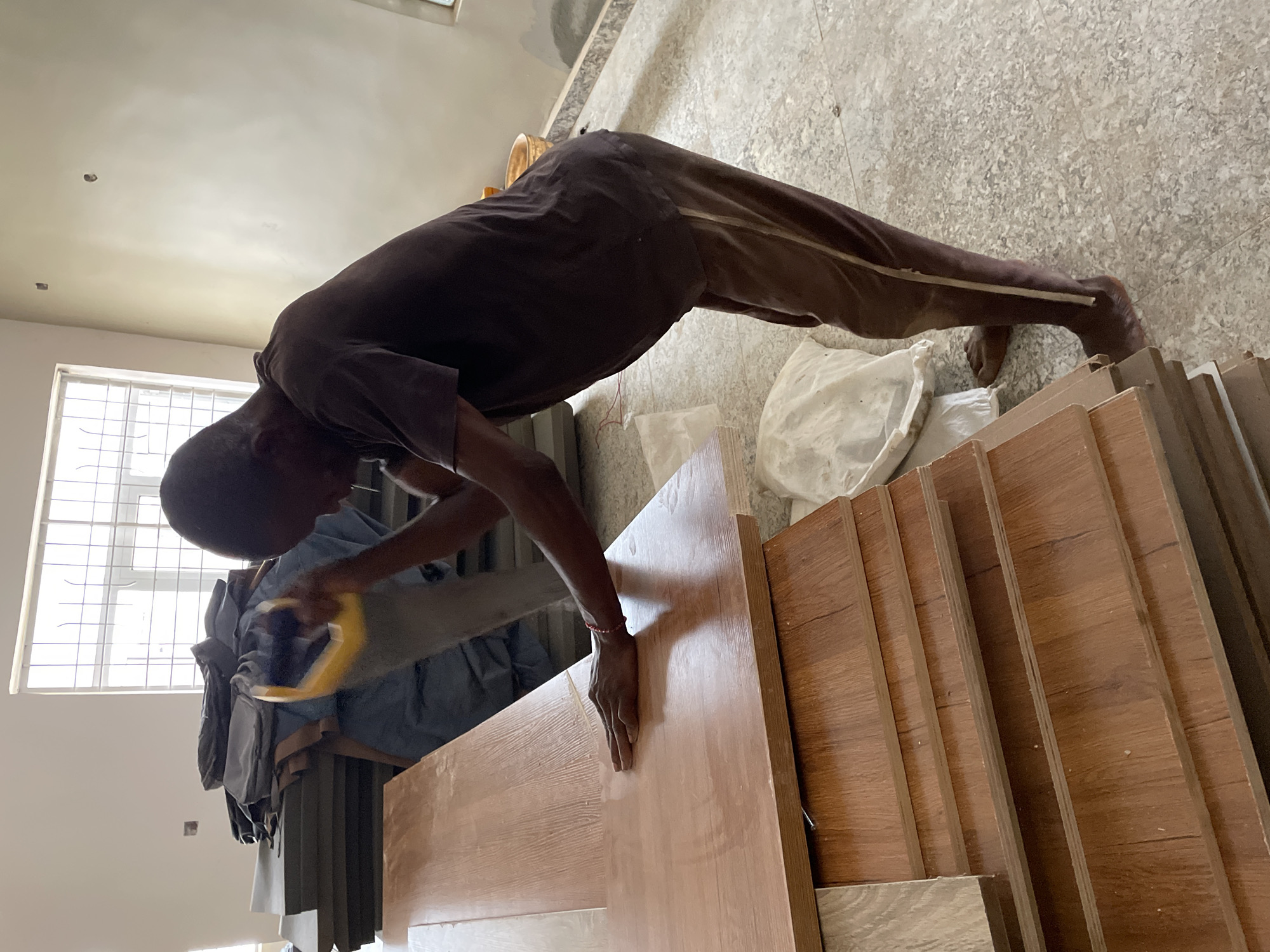 In Jos, Nigeria, a man is sanding a piece of light-colored wood, which is part of a stack of similar wood pieces with a darker wood grain finish. He is wearing a dark t-shirt and dark pants, and standing barefoot on a light speckled floor. A white bag is near the stack of wood, and a window is visible in the background, letting in natural light. The room appears to be under construction or renovation, as indicated by the unfinished walls and ceiling.