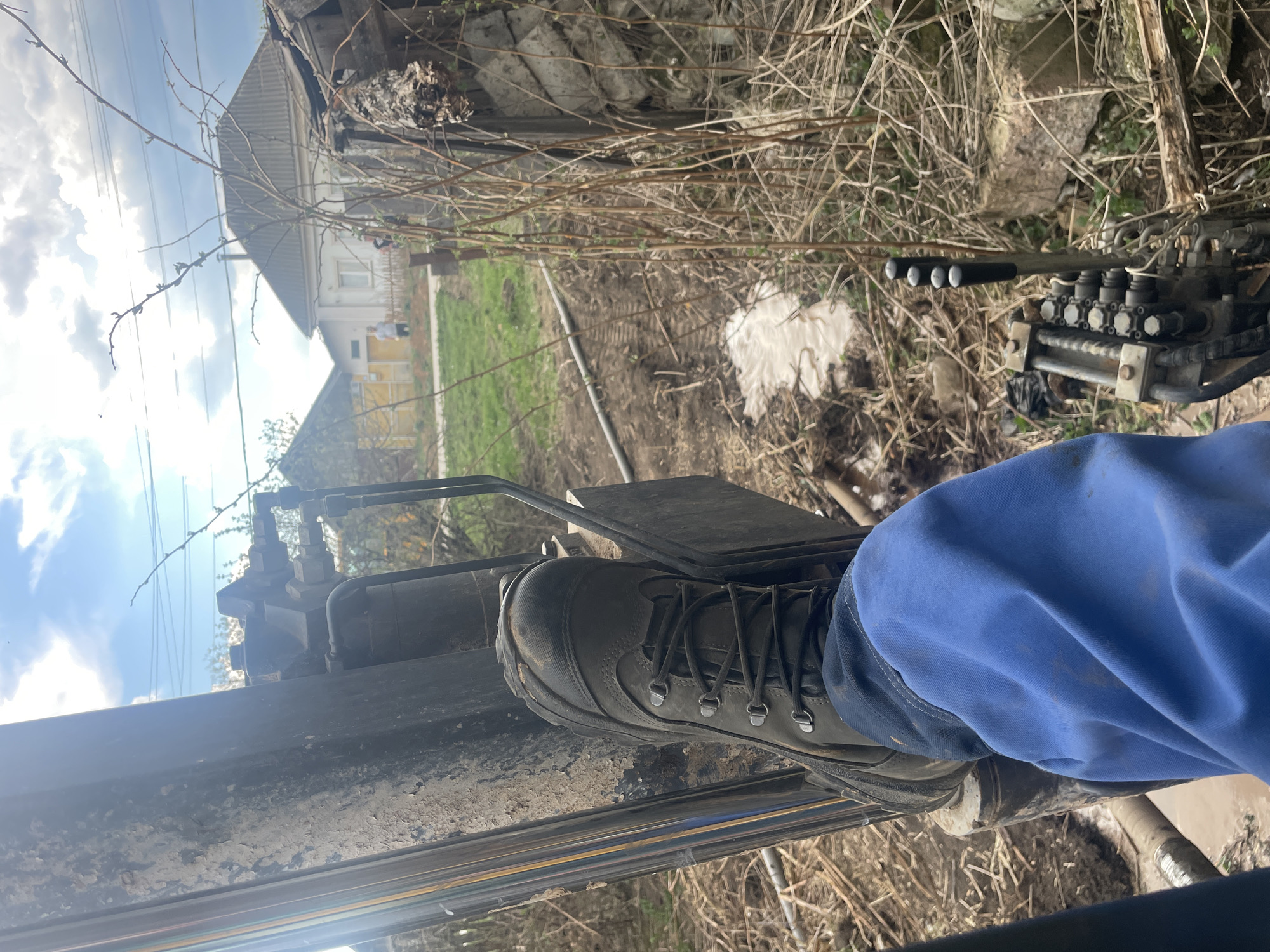 The image shows a close-up view from inside a piece of heavy machinery, possibly a digger or tractor. The viewer is positioned within the cab, looking out. In the foreground, a person's foot, clad in a work boot and blue pants, rests on a metal pedal or footrest.
Outside the machinery, the environment appears to be a rural or suburban setting. There is a patch of grassy land, some tangled dry vegetation, and a building in the background. The building is light-colored and has a gabled roof. Power lines stretch across the upper left of the frame, against a sky with blue and white clouds.
The location is Поедуги, Russia.