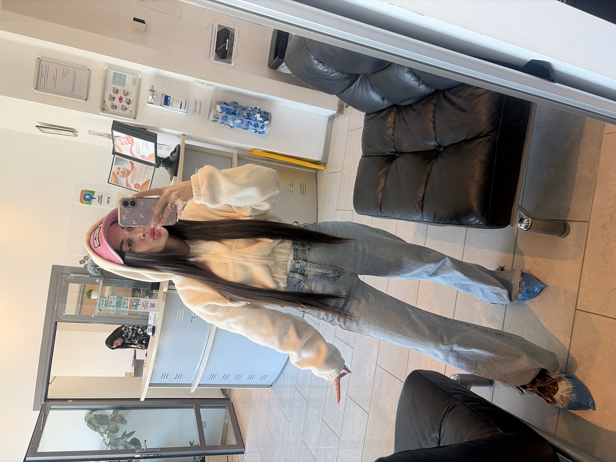 A woman captures a mirror selfie in what appears to be a waiting area or reception of a clinic or salon in Saint Petersburg, Russia. She is dressed in a cream-colored, fluffy hooded jacket, light blue wide-leg jeans, a pink baseball cap, and blue disposable shoe covers over her footwear. Her long, dark hair cascades over her shoulders as she holds a smartphone with a floral-patterned case up to the mirror, looking at her reflection with a slight pout.
The indoor setting features white walls, a light-tiled floor, and a large black faux leather sofa or bench. On the wall behind her, several framed documents and posters are displayed, including one labeled "ИНФОРМАЦИЯ" (Information). Nearby, a dispenser, likely for hand sanitizer, and a wall-mounted rack containing a roll of blue "БАХИЛЫ" (shoe covers) are visible, emphasizing a hygienic environment. Brochures featuring images of faces, possibly related to medical or cosmetic services, are arranged on a display stand. A large mirror in the background reflects a glimpse of another person and some greenery. The bright lighting suggests it is daytime.