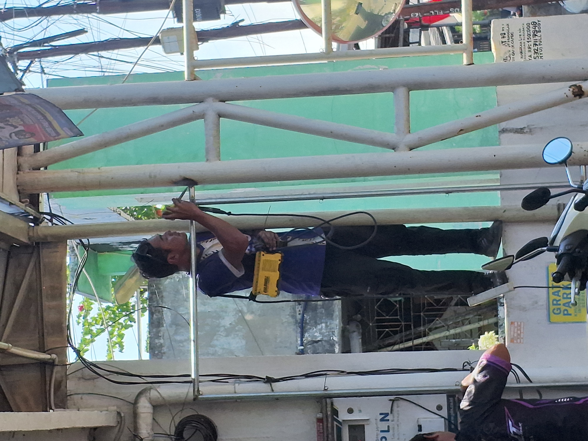A man in a blue shirt and black pants is standing on a metal structure, holding a yellow device. He appears to be working, possibly on electrical wiring or equipment. Wires and conduits are visible around him, suggesting an outdoor or industrial setting. In the background, there are buildings and vegetation. A sign with Indonesian text is partially visible, possibly indicating a location in Surabaya, Indonesia. Another person is seen in the foreground, also wearing dark clothing and looking towards the man. A motorcycle is parked nearby. The overall scene suggests a street-level view of an urban environment in Indonesia.