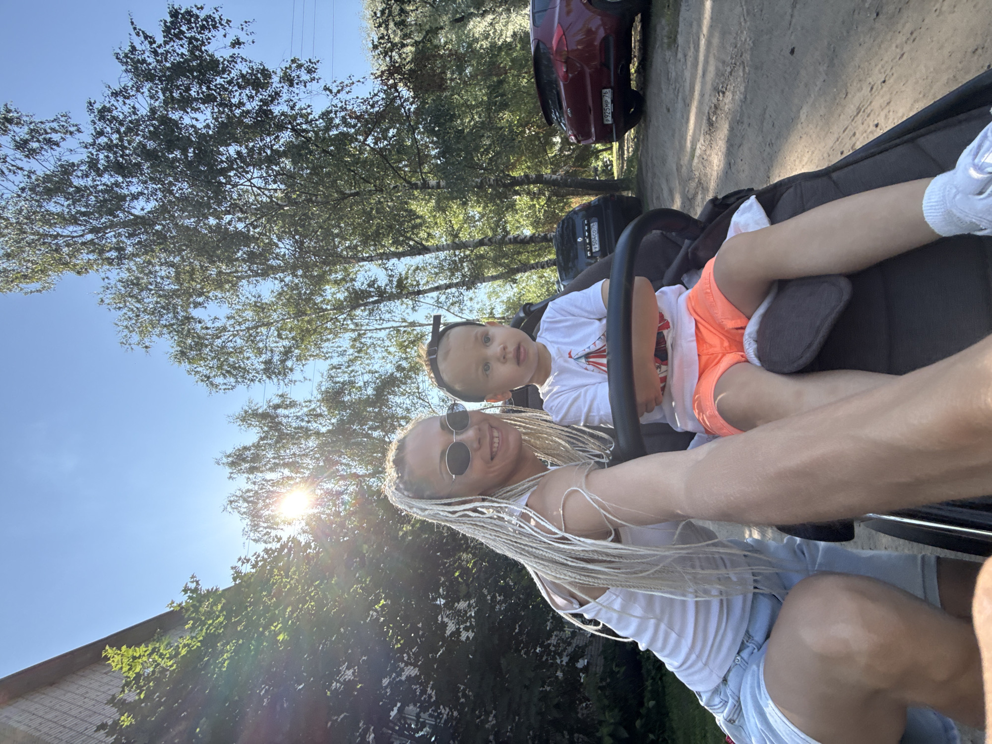 In Rusanovo, Russia, on a sunny day, a woman with blonde braided hair, wearing a white tank top and denim shorts, is seen from the chest up, smiling at the camera. She is next to a child in a stroller. The child, wearing a white t-shirt with an orange and red graphic and orange shorts, looks forward with a neutral expression. The stroller is a dark grey color. In the background, there are trees and a dirt road. To the right of the stroller, two cars are parked: a black sedan with a license plate starting with "Y504", and a red hatchback with a license plate starting with "P925MP 76". A building with a brick facade is visible in the upper left corner of the image, partially obscured by trees. The sun is shining brightly, creating lens flare in the upper left quadrant.