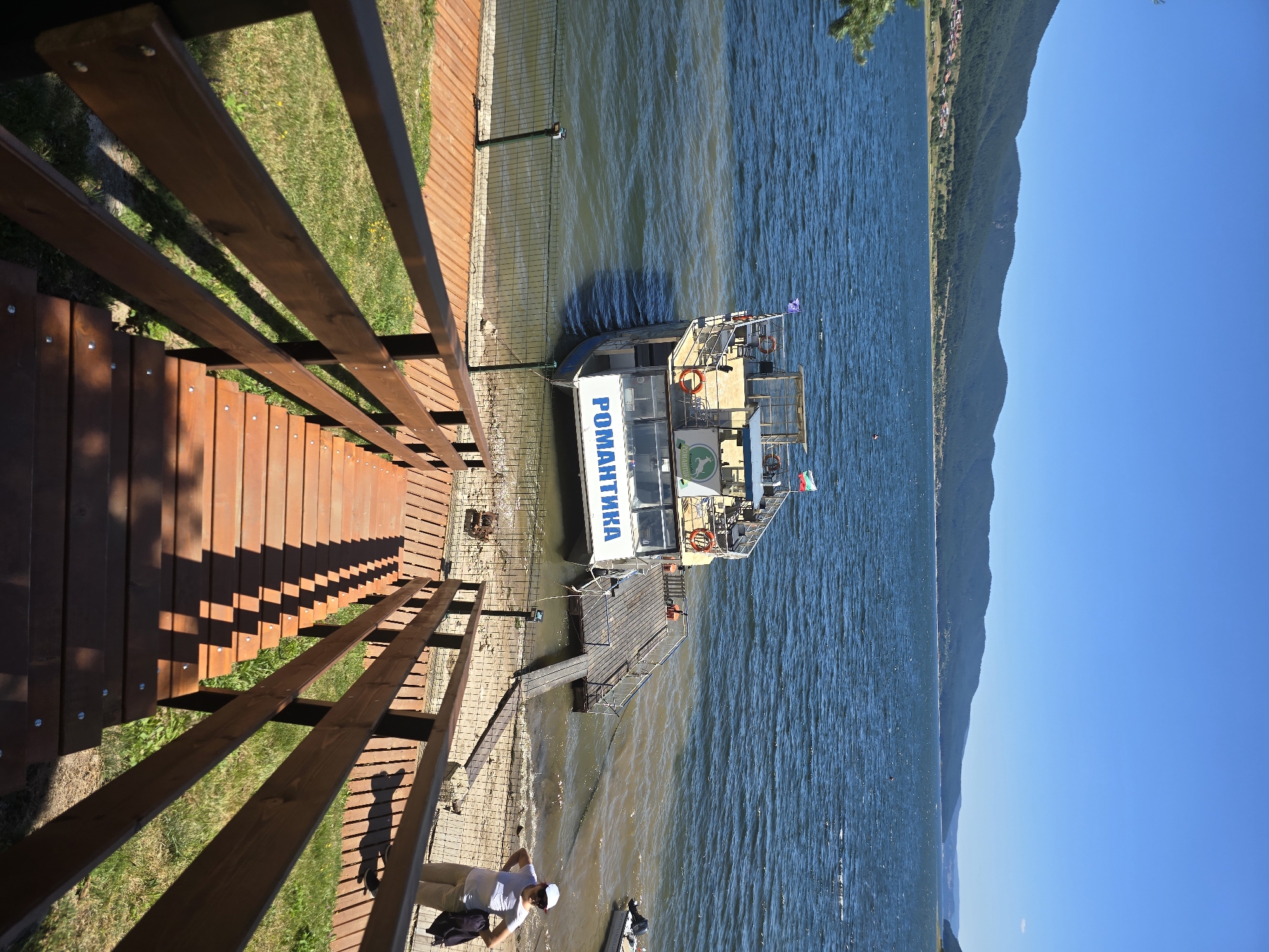 A wide shot captures a sun-drenched lakeside scene with a wooden staircase in the foreground leading down towards the water. A ferryboat named "РОМАНТИКА" is docked at a wooden pier. A woman in a white cap and light-colored pants stands on the shore to the right of the pier. The lake is blue, with a line of green hills in the background, and a clear blue sky above. The scene appears to be in a tourist area.