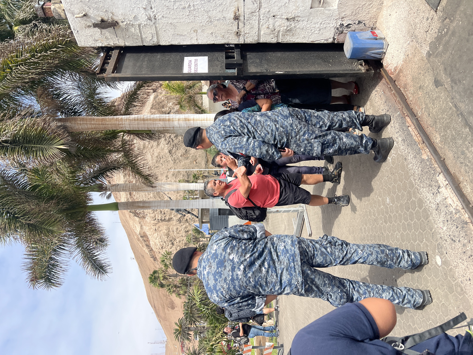 The scene depicts a gathering of people and uniformed personnel near what appears to be an entrance or gate. Two people in blue camouflage uniforms and black berets are standing in front of the gate. The gate is black and has a sign visible on it. A man in a red shirt and black shorts is standing nearby, possibly interacting with one of the uniformed individuals. Several other individuals are visible in the background.
The environment includes palm trees and a sandy cliff or hillside. The location is believed to be in Arica, Chile. The surface underfoot appears to be a paved or concrete area.