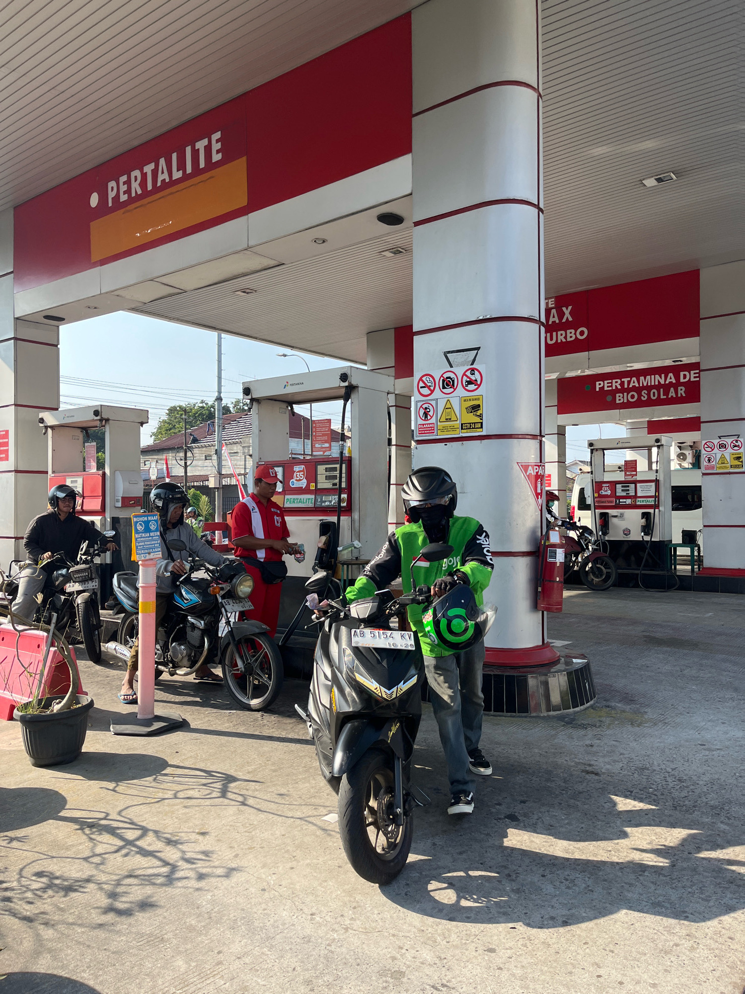 This is a photograph of a Pertamina gas station in Yogyakarta, Indonesia. In the foreground, two men on motorcycles are waiting to refuel. One man, wearing a green jacket and black helmet, is facing the camera and holding his helmet. The other man, wearing a black jacket and helmet, is seated on his motorcycle and looking to the left. Behind them, a gas station attendant in a red uniform is attending to a motorcycle.
The gas station has red and white branding, with signs indicating "PERTALITE" and "PERTAMINA DE BIO SOLAR." The environment appears to be an outdoor, open-air space with a concrete ground. The lighting suggests it is daytime, with shadows cast on the ground from the overhead structures and the people. In the background, other gas pumps and vehicles are visible, as well as some buildings and trees. The overall scene depicts a common daily activity of refueling vehicles at a gas station in Indonesia.