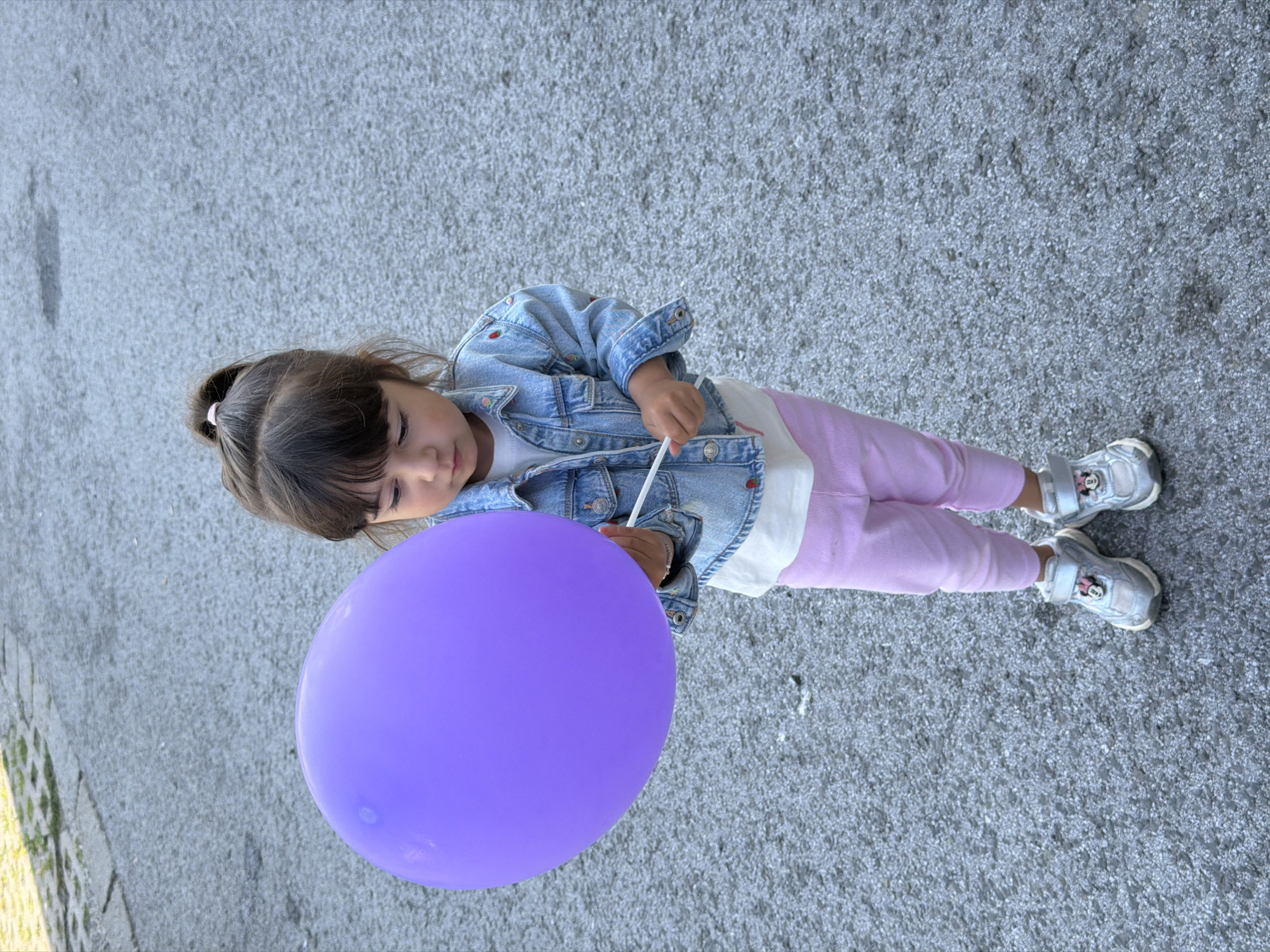 A young girl stands on a grey, coarse-textured paved surface in Burgas, Bulgaria. She has dark hair tied in a high ponytail and is wearing a light blue denim jacket with small red embroidered accents, a white t-shirt, light pink leggings, and silver sneakers with Minnie Mouse emblems on the Velcro straps. She holds a spherical, vibrant purple balloon by a white stick in her hands, looking downwards at the balloon. In the upper left background, a lighter paved area with minimal green vegetation is partially visible.