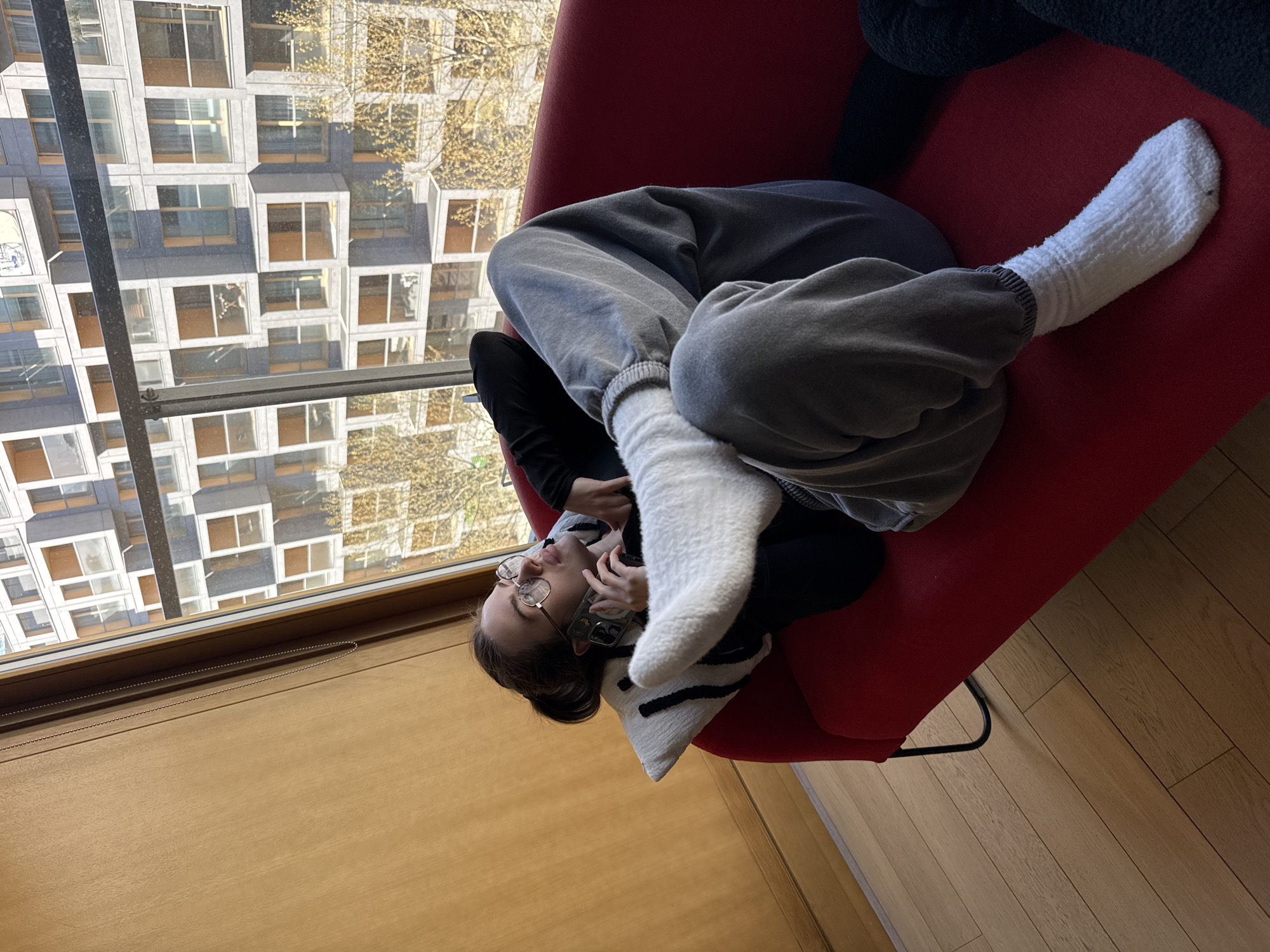 An indoor scene captured from a high angle shows a person lying comfortably on a bright red armchair next to a large window, overlooking a cityscape in Warsaw, Poland. The person, a young adult with dark hair and glasses, is positioned with their head near the bottom of the frame, appearing upside down relative to the window view. They are dressed in grey sweatpants, a dark top, and thick white fuzzy socks, with their legs bent and feet propped up. A white pillow with black stripes is visible under their head. The person is holding a smartphone to their left ear, seemingly engaged in a phone call.
The window reveals a view of modern, multi-story buildings with numerous square and rectangular windows, reflecting the bright daylight. The architecture suggests a contemporary urban environment. There are also bare tree branches visible against the buildings, indicating a late autumn or winter season. Sunlight casts clear shadows on the buildings, suggesting a sunny day. The room itself features a wooden floor with visible planks and a window frame with a blind control chain. The overall atmosphere appears relaxed and casual. There is no visible text in the image.