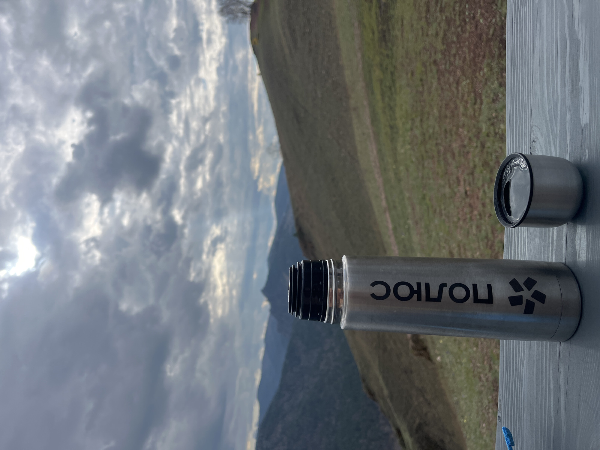 The image captures a landscape scene with a water bottle and its cap in the foreground, positioned on a wooden surface. The background reveals a mountainous terrain under a cloudy sky. The mountains, covered in green vegetation, extend towards the horizon, partially obscured by the atmospheric perspective of the sky. The bottle has the word "ПОЛЮС" on it. The bottle and cup seem to be on a wooden table. The location is unspecified in the prompt and therefore unknown.