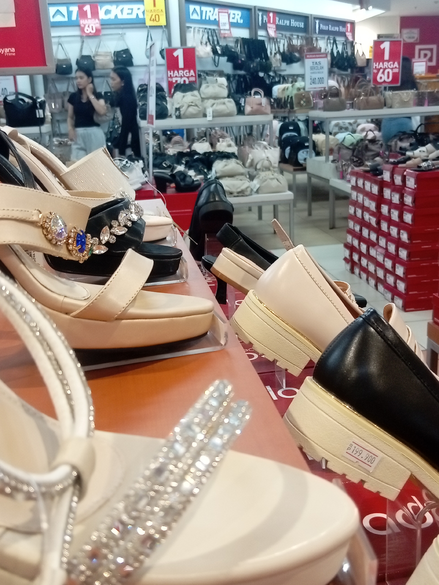 The image shows a retail display of women's footwear, primarily high-heeled sandals and pumps. The shoes are arranged on shelves and a counter, with some in their boxes stacked on the right side of the frame. The setting appears to be a department store or shoe shop, likely located in Batam, Indonesia, given the pricing labels which show "Rp" (Indonesian Rupiah). There are two women visible in the background, one in a black shirt and light-colored pants, and the other in a black top, both appearing to be browsing or working in the store. The lighting suggests an indoor environment. One pair of beige high-heeled sandals in the foreground is decorated with rhinestones and a blue jewel accent. Another pair of black pumps in the mid-ground also features rhinestone embellishments. A price tag on a pair of black platform shoes reads "Rp 199.900".