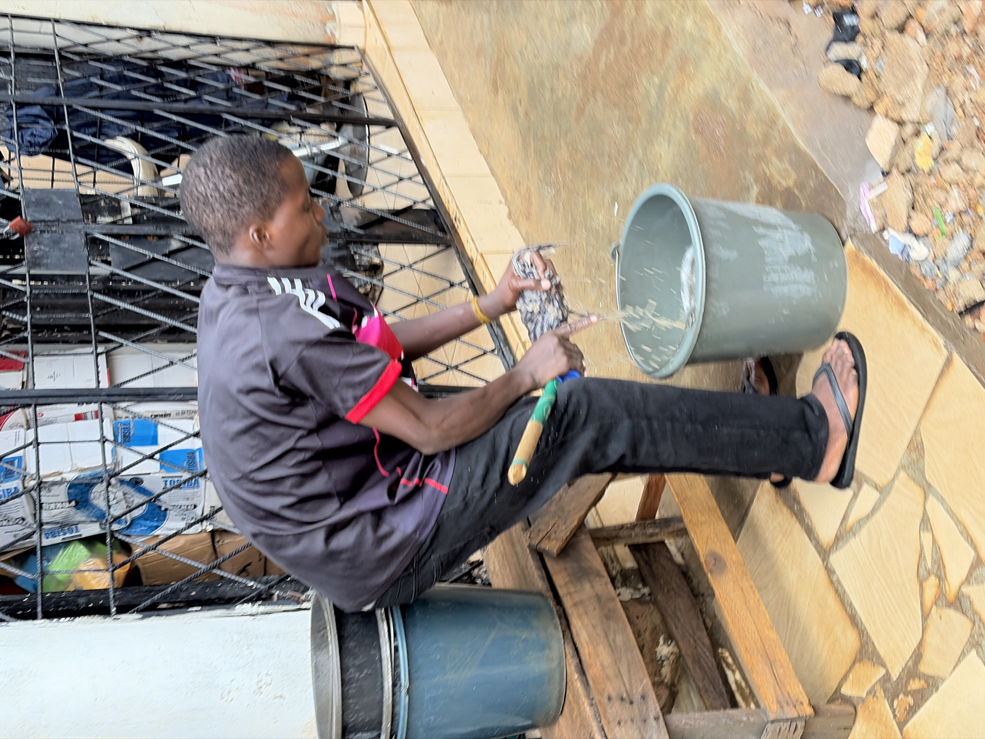 A young person wearing a black and pink athletic shirt and black pants is sitting on a wooden structure, outdoors, near a building with a barred window. The person is holding a bird in their hands and appears to be tending to it. A grey bucket is positioned next to them, and another blue bucket is below the wooden structure. The ground is paved with irregular, light-colored tiles. Debris and construction materials are visible in the background. The lighting suggests it is daytime. The scene appears to be in Jos, Nigeria.