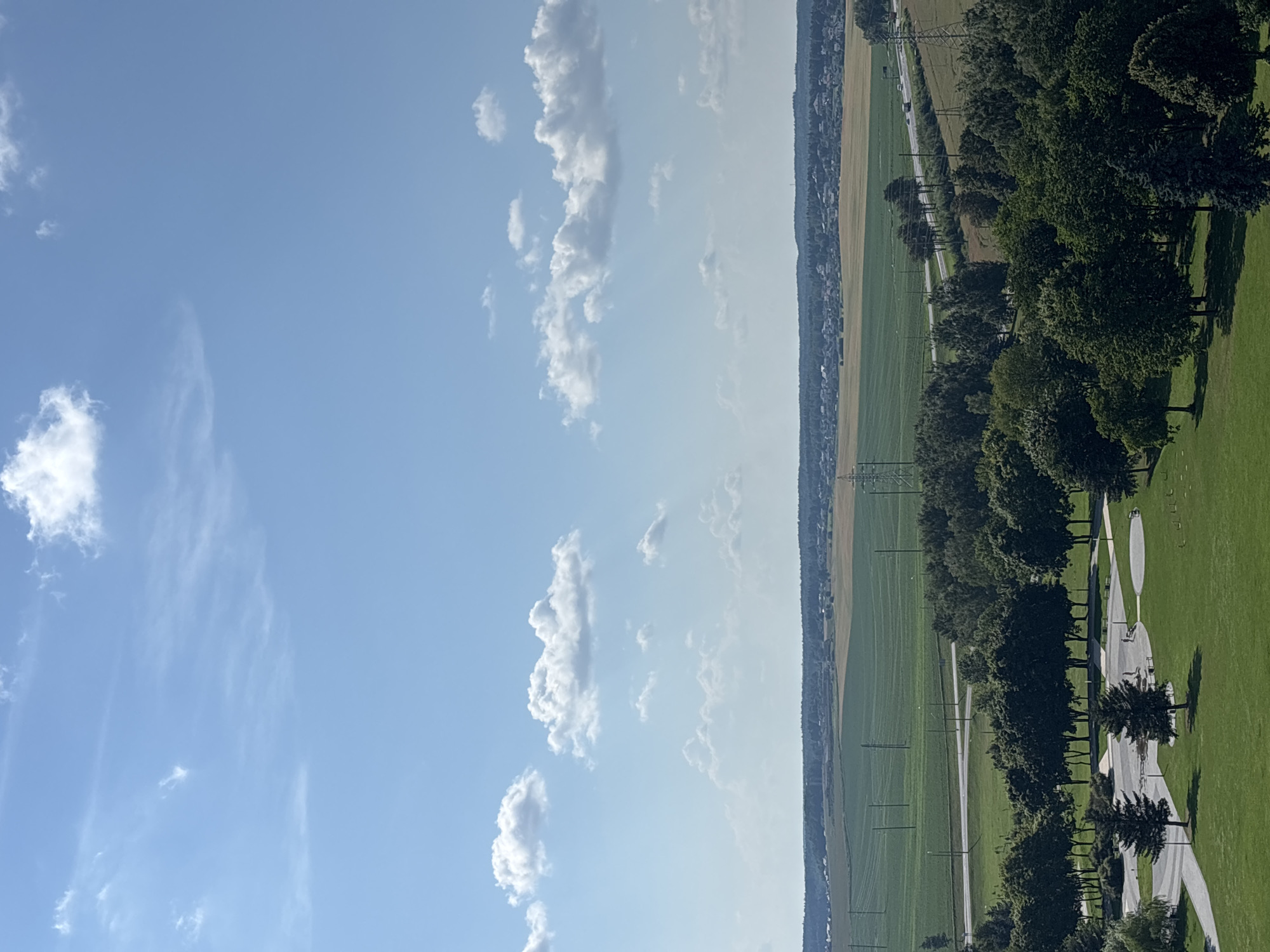 The image depicts a panoramic view of a park area leading to agricultural fields and a highway, under a bright blue sky with scattered white clouds. In the foreground, a well-maintained park features manicured green lawns, paved pathways, benches, and several trees, including evergreen species. Further into the scene, a line of deciduous trees separates the park from a paved road with sparse traffic. Beyond the road, vast green agricultural fields stretch towards a distant treeline and a settlement. The sky is clear and sunny, with wispy clouds creating a dynamic contrast against the blue. The overall impression is one of a serene, rural landscape on a clear day. The location is identified as being within the Ozyaritska-Slabadski rural council in Belarus.