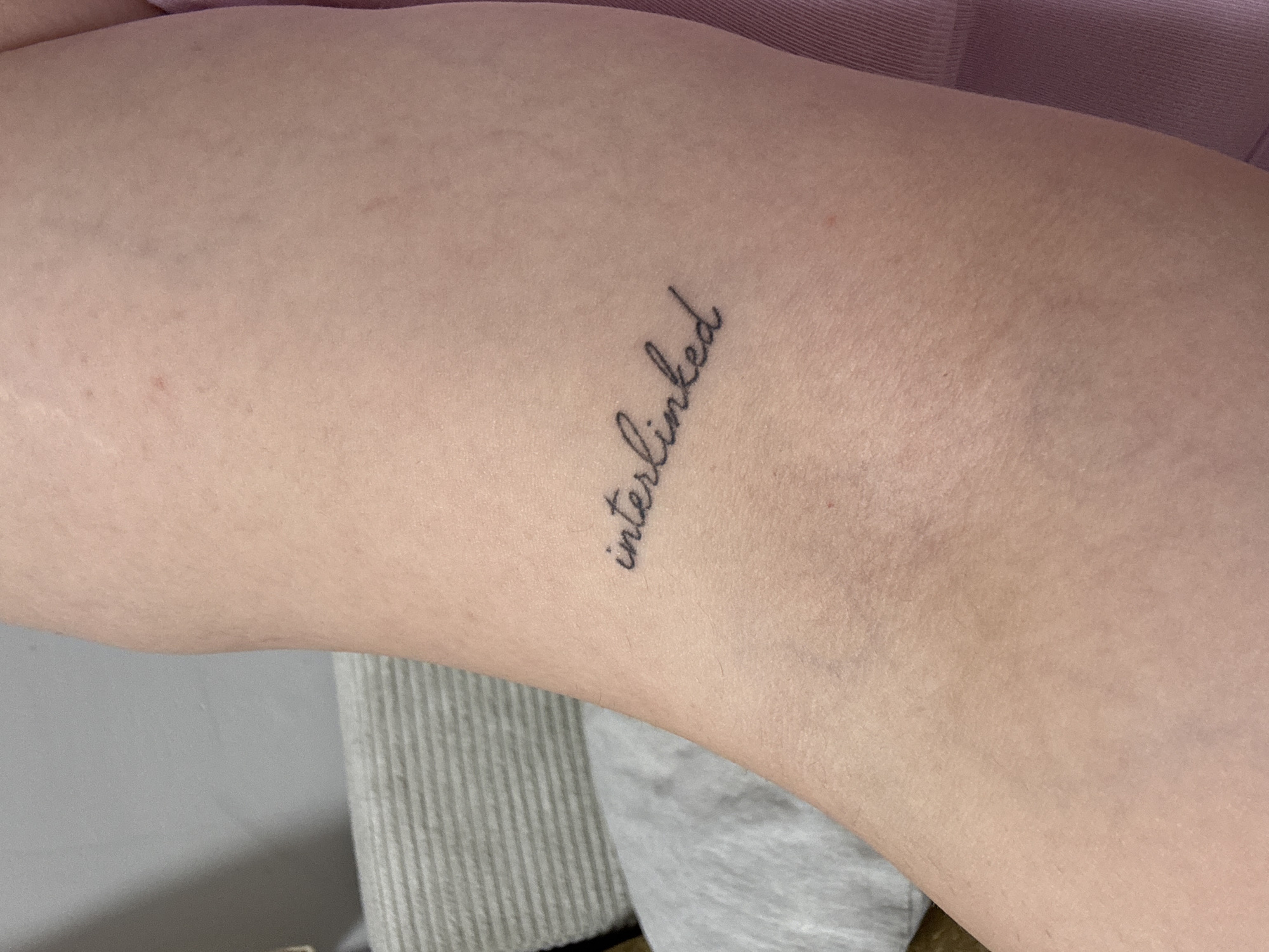 The image shows a close-up of a person's arm or leg with a tattoo that reads "interlinked" in cursive script. The skin tone is fair. The background is blurred and includes a gray textured surface, possibly fabric or upholstery, and a hint of a pink garment. The image does not contain information suggesting a specific location in Zlatitsa, Bulgaria.