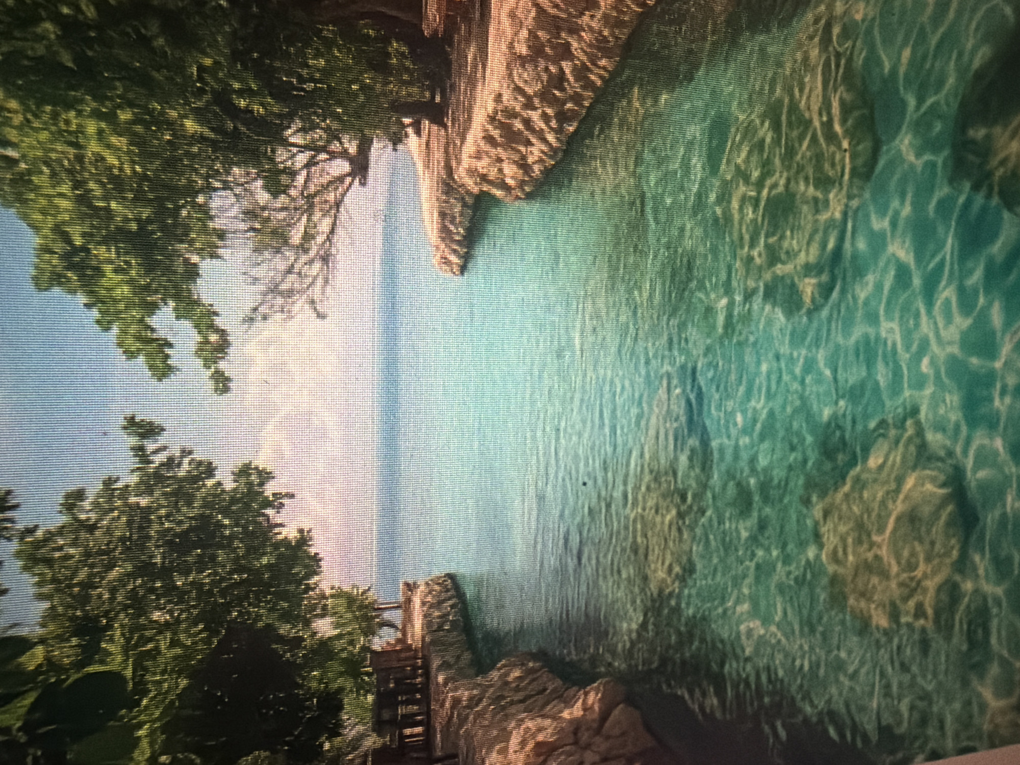The image depicts a serene tropical inlet with crystal-clear turquoise water. The water is shallow enough to reveal submerged rocks and the sandy bottom. Lush green trees overhang both sides of the inlet, casting dappled shadows on the rocky shorelines. On the left side, a small wooden seating area with tables is visible on a stone-lined embankment. The sky is a pale blue with soft white clouds. The scene conveys a sense of tranquility and natural beauty. The location is described as Baan Mae Nam, Thailand.
