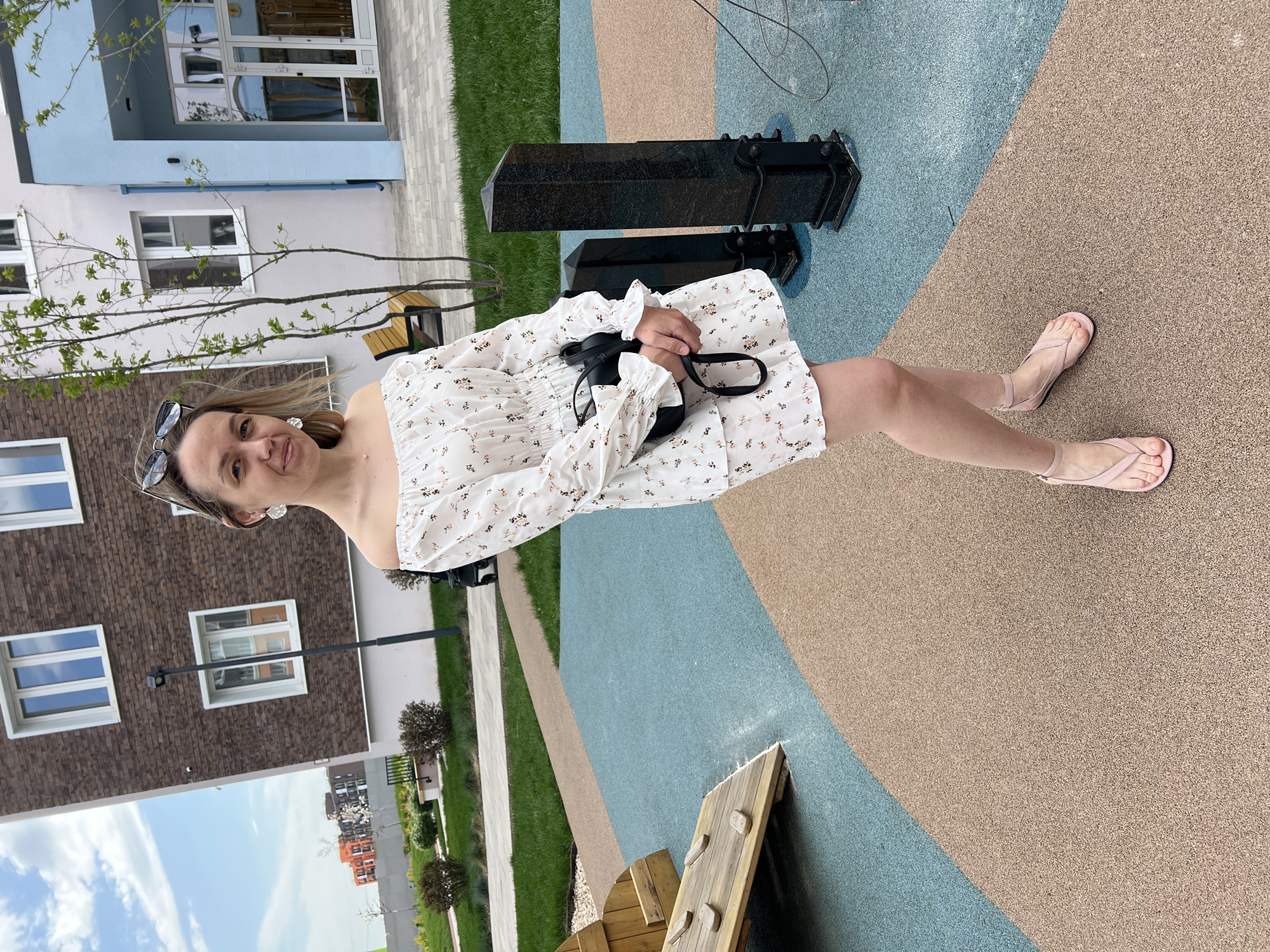The image depicts a woman in a white floral dress and pink sandals standing on a playground surface with tan and blue sections. She is holding a black purse. In the background are apartment buildings, green lawns, and a small black object possibly related to play equipment. The location is Pervomaysky, Russia.
