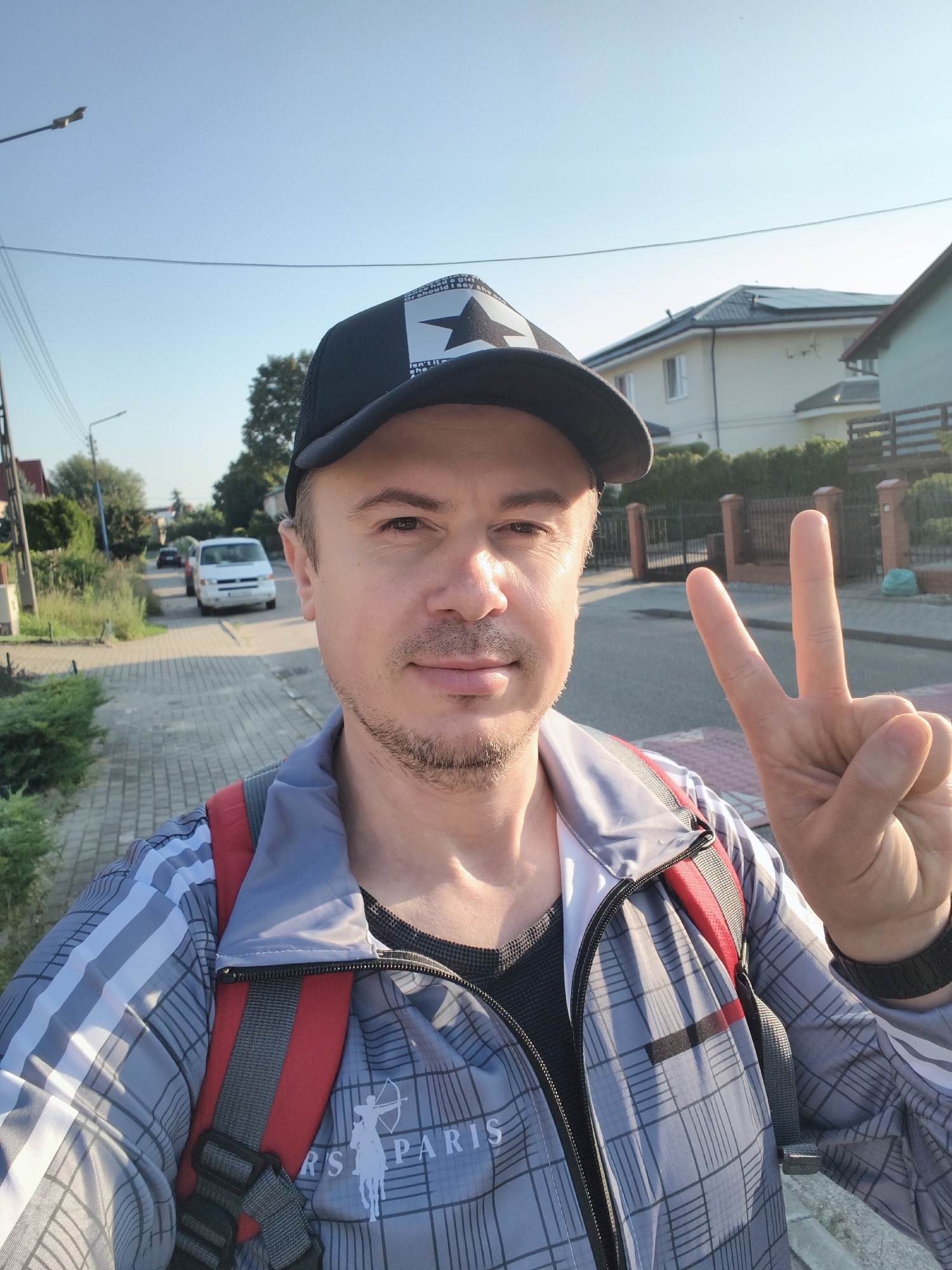 A man wearing a black baseball cap and a plaid jacket over a red backpack is taking a selfie outdoors in a suburban area. He is holding up two fingers in a peace sign. In the background, there is a paved walkway, houses with solar panels on their roofs, a fence, and parked cars. The overall lighting suggests it is daytime. The location has been identified as Tczew, Poland.