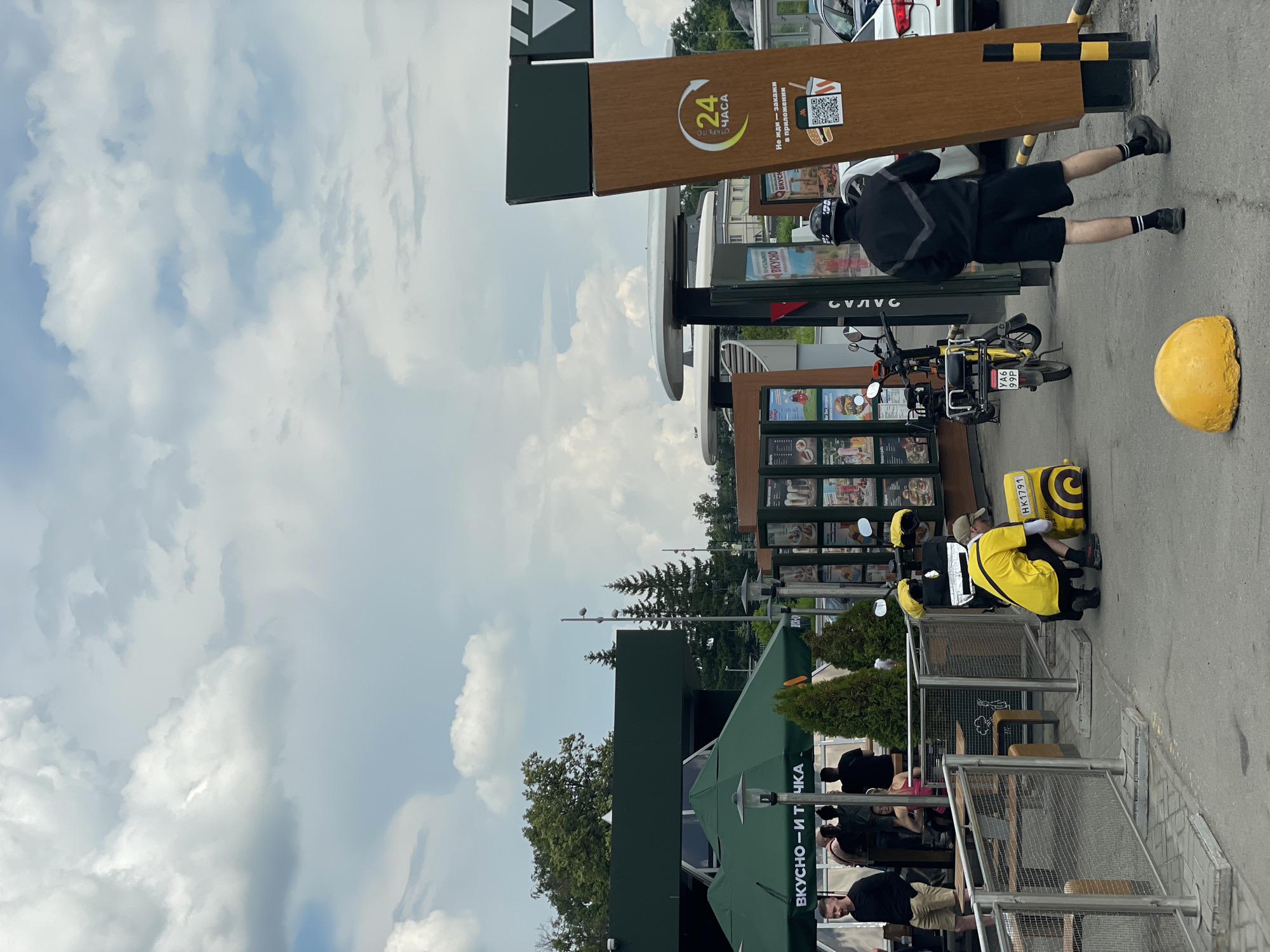 In Kazan, Russia, outside a McDonald's restaurant, two delivery drivers in yellow uniforms are preparing food orders. One driver, wearing a helmet, is crouched next to a yellow delivery backpack placed on the ground, while the other stands nearby. A yellow electric scooter, also equipped with a delivery box, is parked behind the crouching driver. In the background, customers are visible sitting at outdoor tables under green awnings. The scene takes place under a partly cloudy sky, with trees and buildings forming the backdrop. The McDonald's features a drive-thru service with a prominent sign indicating "24 HOURS." A white car is parked to the right of the drive-thru lane.