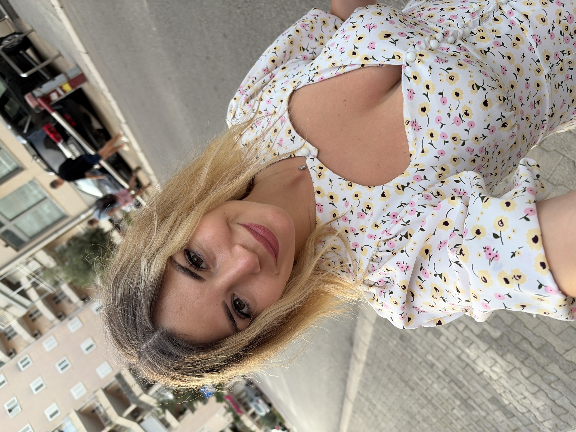 The image shows a woman with blonde hair taking a selfie outdoors on a street in Budva, Montenegro. She is wearing a white dress with a floral print of pink and yellow flowers. The dress has short puff sleeves and a cutout detail at the neckline. She is smiling and looking at the camera.
In the background, there are apartment buildings with balconies, a street with cars parked along the side, and several other people walking. The street is paved with cobblestones on one side and asphalt on the other. The scene appears to be in a populated urban area.