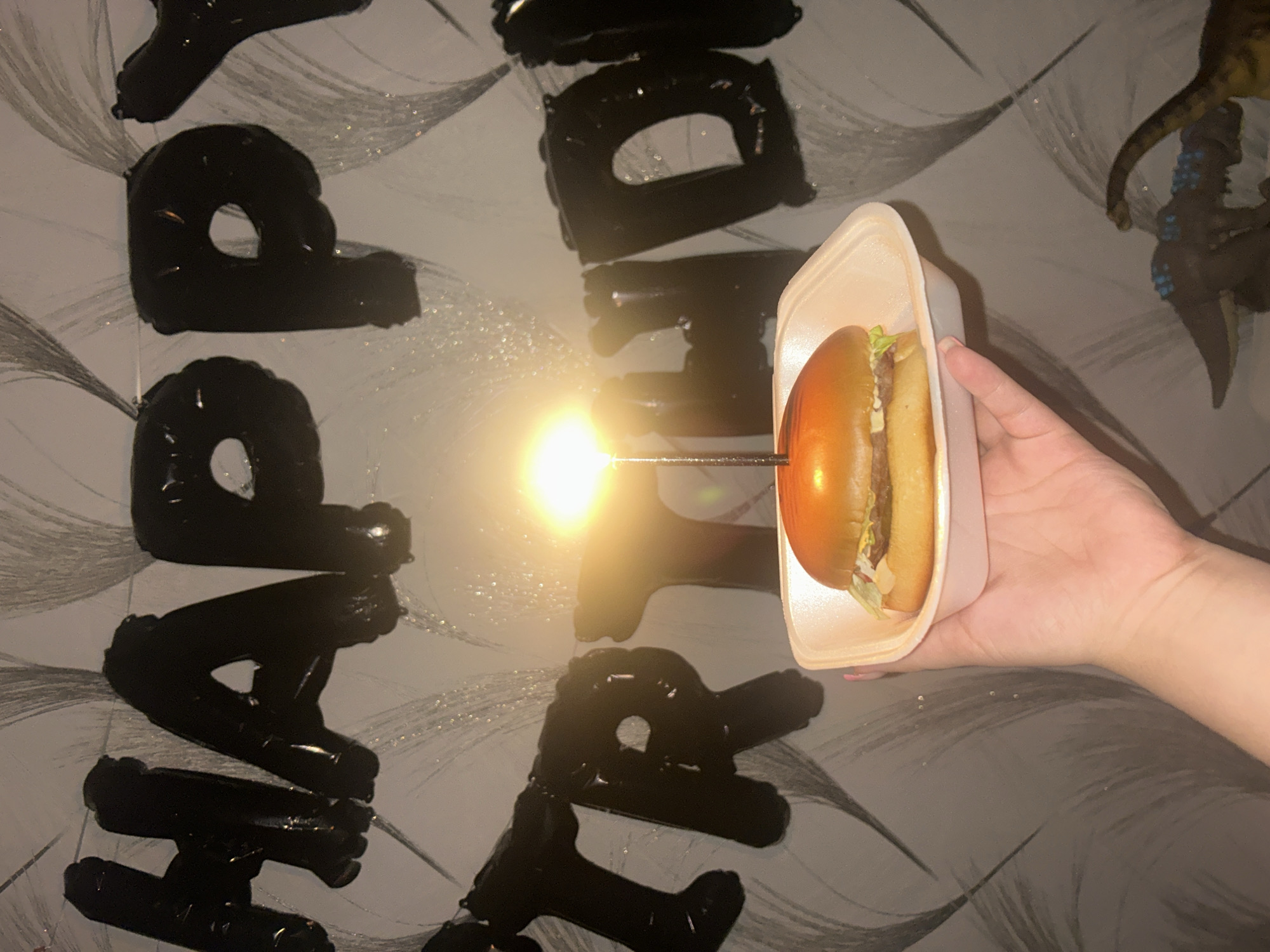 A hand holds a burger in a white container with a lit candle inserted in the bun. The word "HAPPY" is spelled out with black balloons on a light gray wall with a patterned design, suggesting a birthday celebration. The location is Chaikovsky, Russia. A toy dinosaur can be seen in the upper right corner.