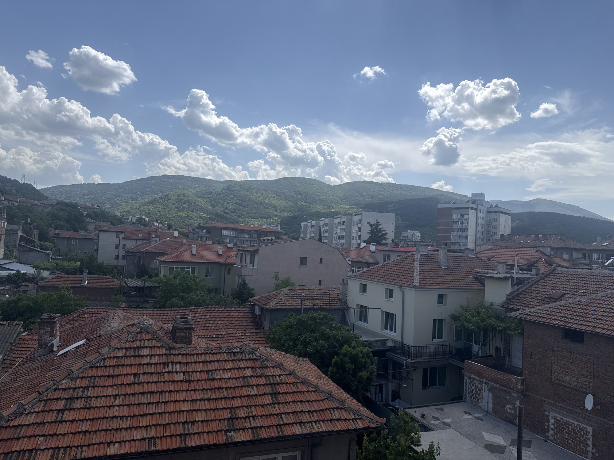 The image depicts an elevated view of a residential area within an unidentified city in an unknown location. Predominantly visible are rows of houses and buildings with distinctive orange tiled roofs. In the background, a range of green hills or mountains forms the horizon line. The sky is blue with scattered white cumulus clouds. The buildings closer to the viewer have varied architectural styles. There are no people visible in the image. The overall environment suggests a sunny day in a built environment set against a natural landscape.