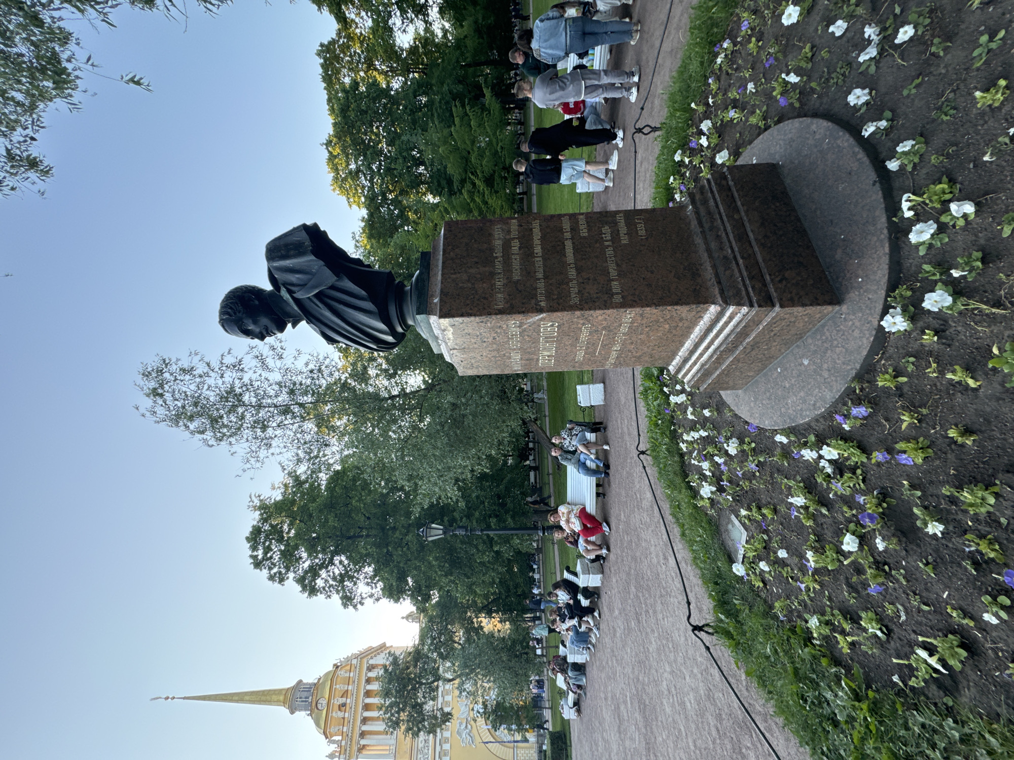 In Saint Petersburg, Russia, a bust of Mikhail Lermontov stands prominently in a park, a granite pedestal bearing an inscription in Russian. Behind the statue, a group of people are seated on park benches, observing the surroundings. In the background, the Admiralty Building, with its distinctive golden spire, looms large against a clear blue sky. Lush green trees and a manicured flower bed with white and purple blooms frame the scene. The lighting suggests it is late afternoon or early evening, casting a soft glow over the park.