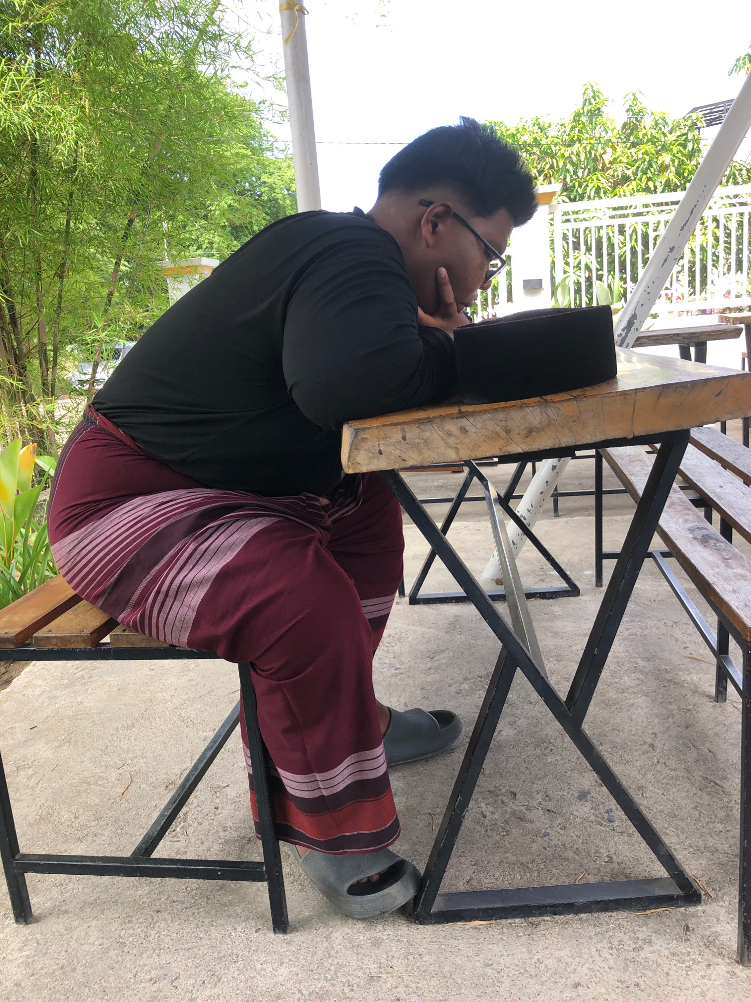 A person wearing a black long-sleeved shirt, dark red sarong with horizontal stripes, and grey slip-on sandals is seated at a wooden table with a black peci on it. The person is looking down at the peci, resting their chin on their hand. The scene appears to be outdoors, with bamboo plants and greenery in the background. The ground is concrete. The table has a metal frame and is located in an outdoor seating area, possibly a cafe or restaurant.