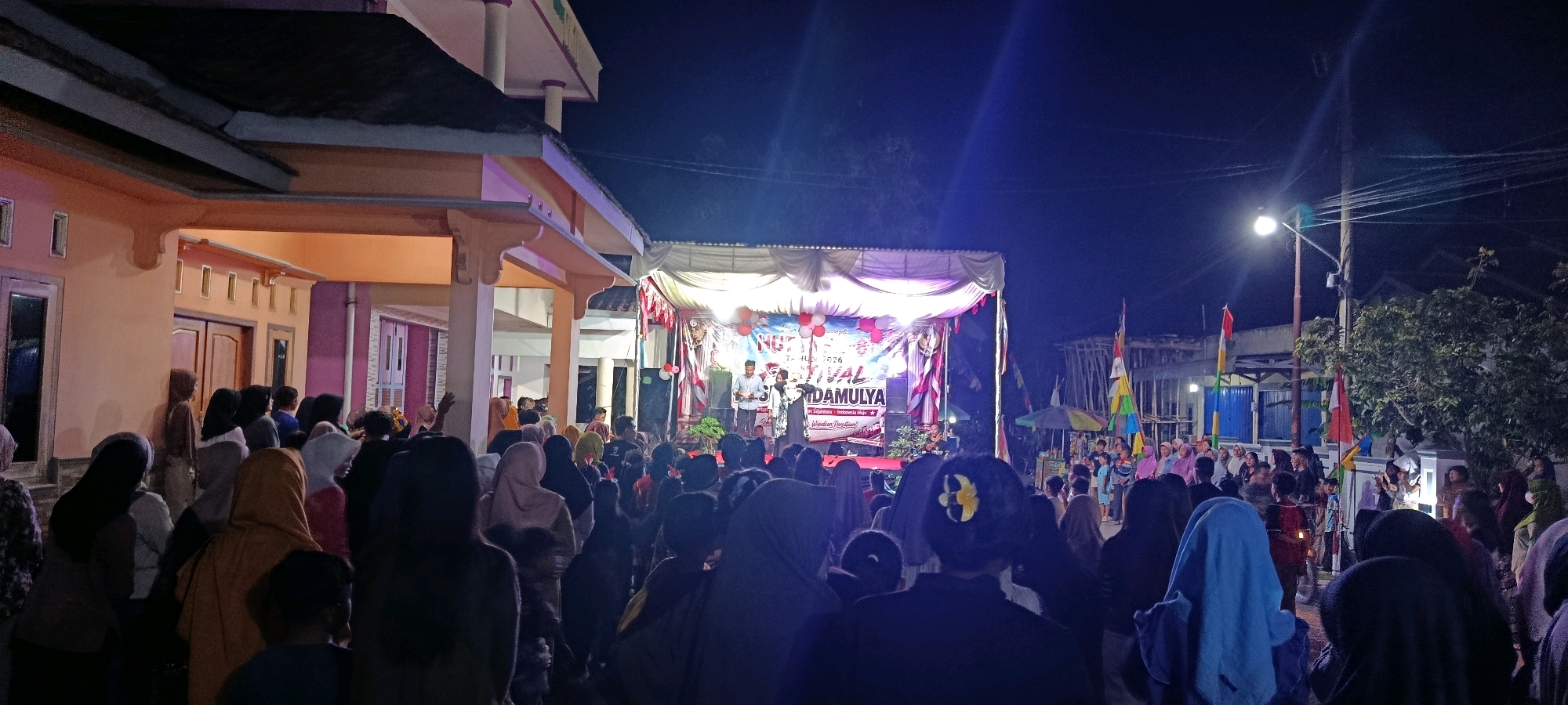 The image depicts an outdoor evening event in the city of Binangun, Indonesia. A stage is set up with a white canopy and decorated with red and white banners, balloons, and what appears to be a floral arrangement. Several people are on the stage, including two individuals holding microphones and another playing a musical instrument. A banner on the stage reads "HUT" and "FESTIVAL DAMULYA" and includes text in Indonesian.
A large crowd of people of various ages is gathered in front of the stage, watching the performance. Many in the crowd are wearing headscarves or hijabs. The event is illuminated by stage lighting and a streetlamp. To the left of the stage is a building with an orange and cream-colored exterior, featuring columns and windows. On the right side of the scene, there are trees, a utility pole with wires, and temporary structures made of wood and what appears to be corrugated metal. Several Indonesian flags are visible, some on poles and some attached to temporary structures. The overall atmosphere suggests a community celebration or festival.