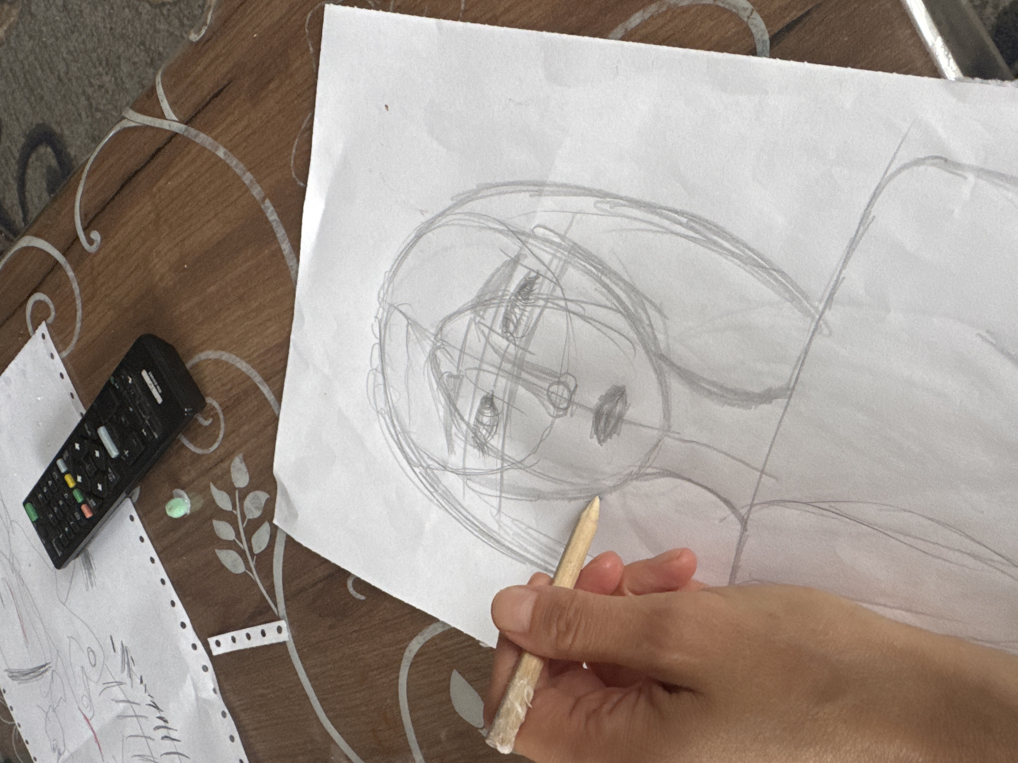 The media file shows a close-up view of a hand holding a pencil over a piece of paper with a sketch of a face. The sketch is a preliminary drawing, with multiple overlapping lines indicating the artist's process of forming the features. The face appears to be drawn in a stylized or abstract manner. To the left of the sketch, a black remote control is visible on a wooden table. The table has a decorative pattern with white swirls and floral motifs. There is also another piece of paper with drawings on it, partially visible in the lower left corner. The overall setting appears to be indoors, with natural light illuminating the scene. No specific time of day or weather conditions are discernible. The emotional tone is neutral, focusing on the act of drawing. No visible text is present in the image. The location context provided is Старое Михайловское, Russia.