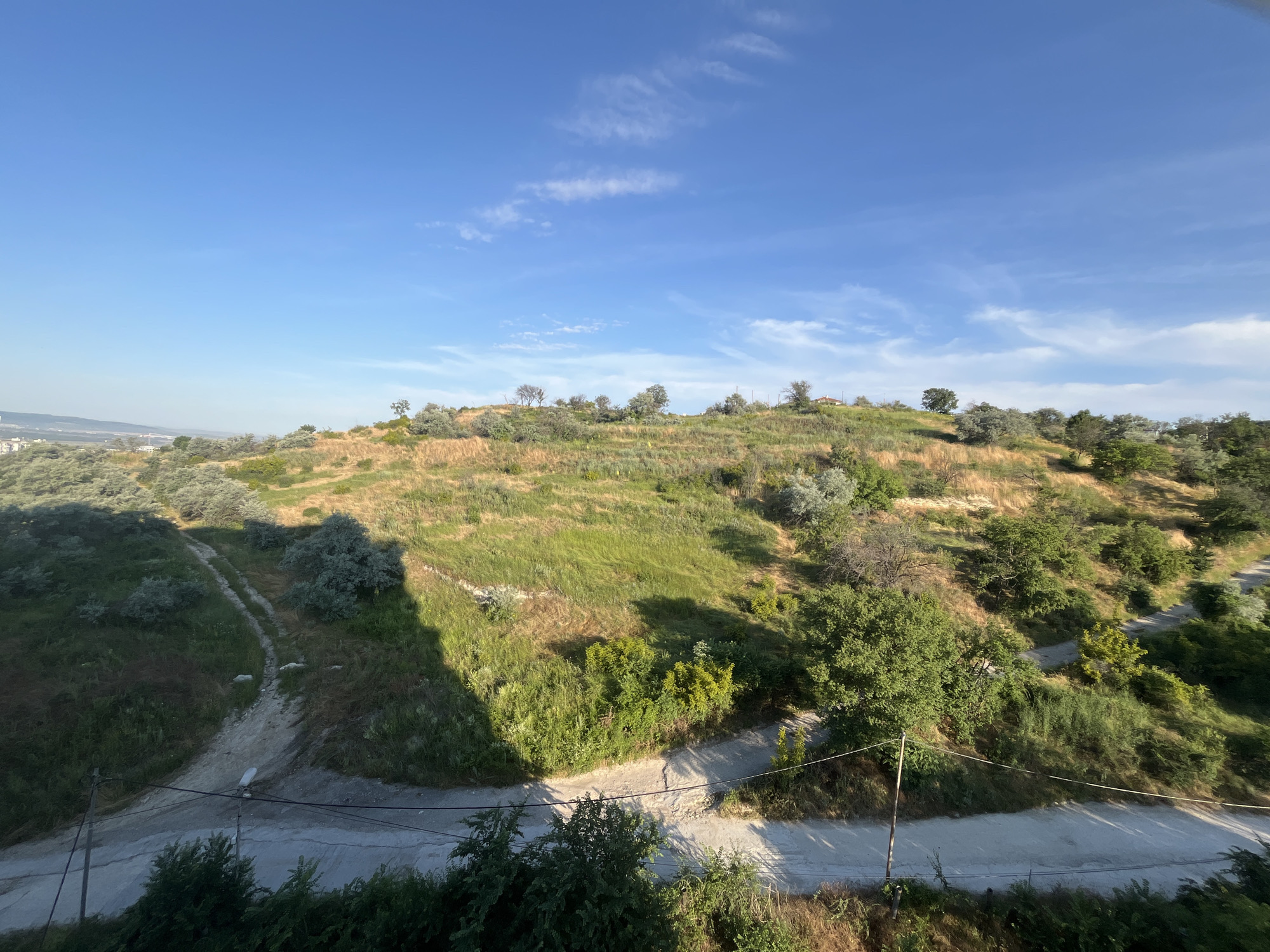 The image captures a sun-drenched hillside in the Vinitsa district of Varna, Bulgaria. A dirt path winds its way up the grassy slope, dotted with sparse trees and shrubs. The sky above is a vibrant blue, with wispy white clouds streaking across it. In the foreground, a concrete road is visible, with power lines and poles running alongside it. The overall impression is one of a rural landscape bathed in the warm light of a summer day.