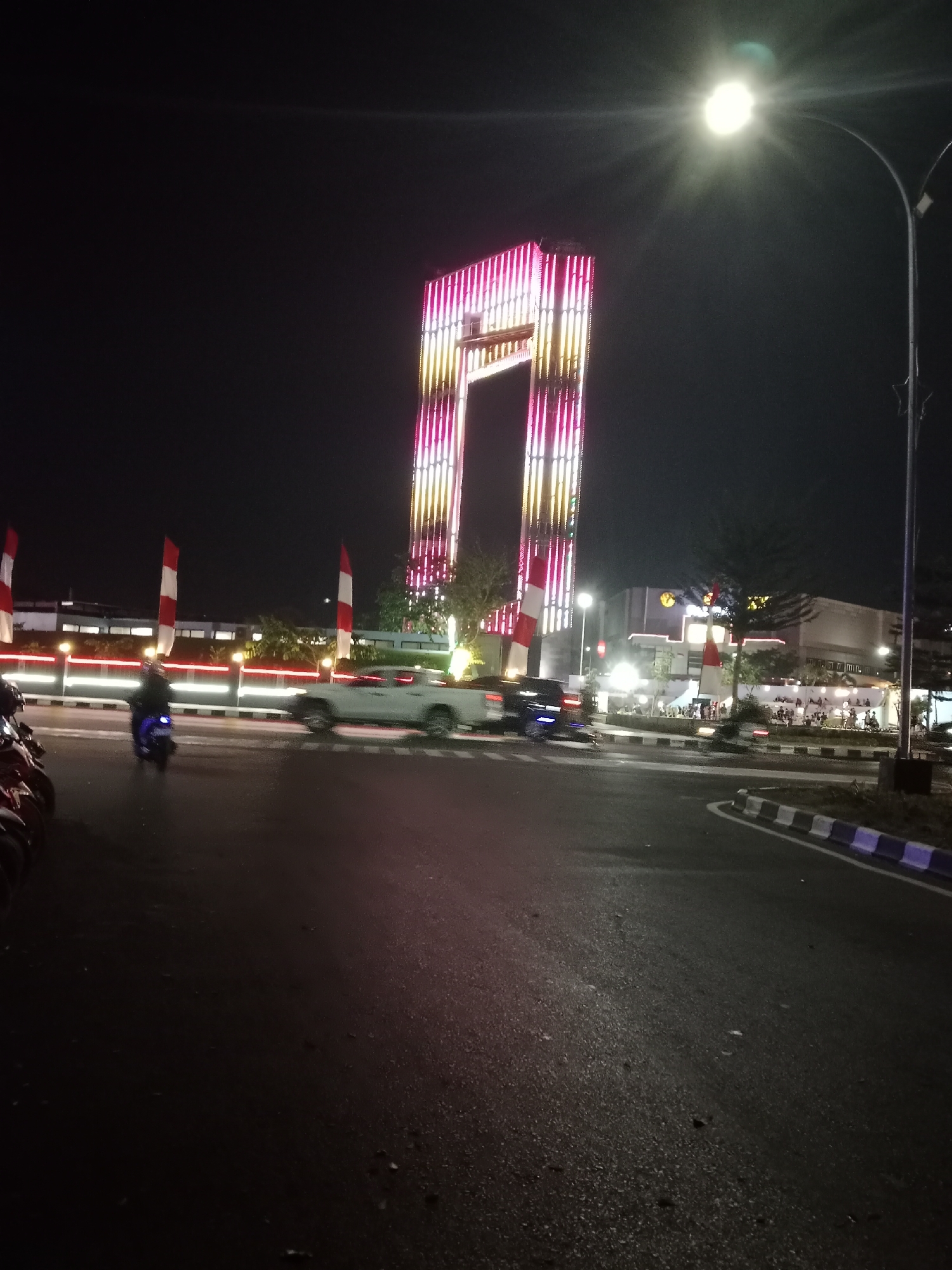 At night, a white pickup truck and a motorcycle are driving on a wet asphalt road. In the background, a tall, brightly lit structure with pink and yellow lights stands out against the dark sky. The structure appears to be a monument or landmark. There are also buildings, trees, and streetlights visible. Red and white flags are planted along the side of the road. The scene suggests an urban setting in Palangka Raya, Indonesia, possibly near a public space or entertainment district, illuminated by artificial lights and the glow of vehicles. The lighting and reflections on the wet road indicate a recent rain or damp conditions.