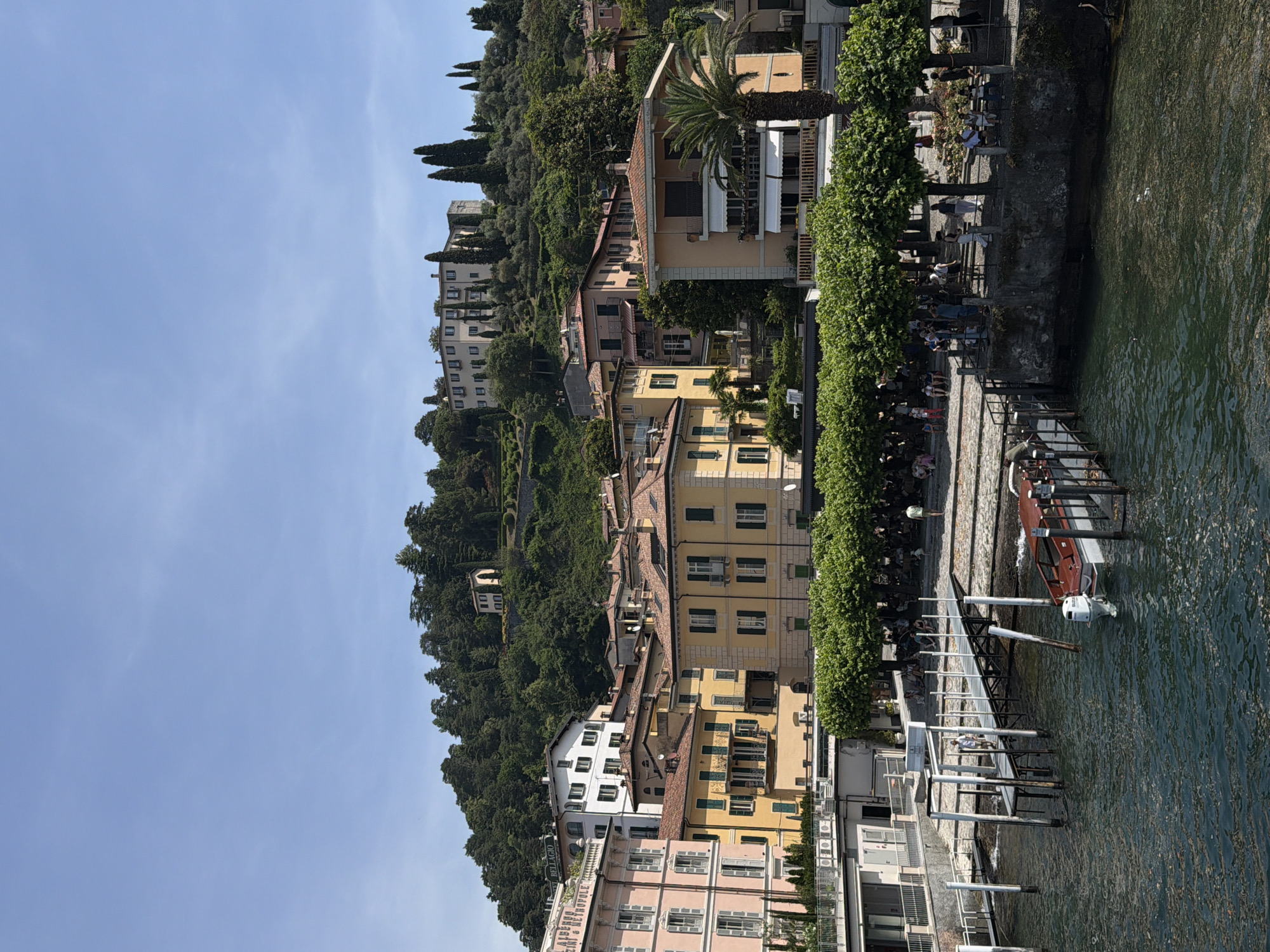 The image depicts a waterfront scene in San Giovanni, Italy, with buildings ascending a tree-covered hill along the water's edge. The buildings are of various colors, including pastel pink, white, and yellow with green shutters. The water's edge has a stone wall and a walkway lined with trees, with people seated on benches. A dock extends into the water, with boats tied to it. A blue sky is visible above.
