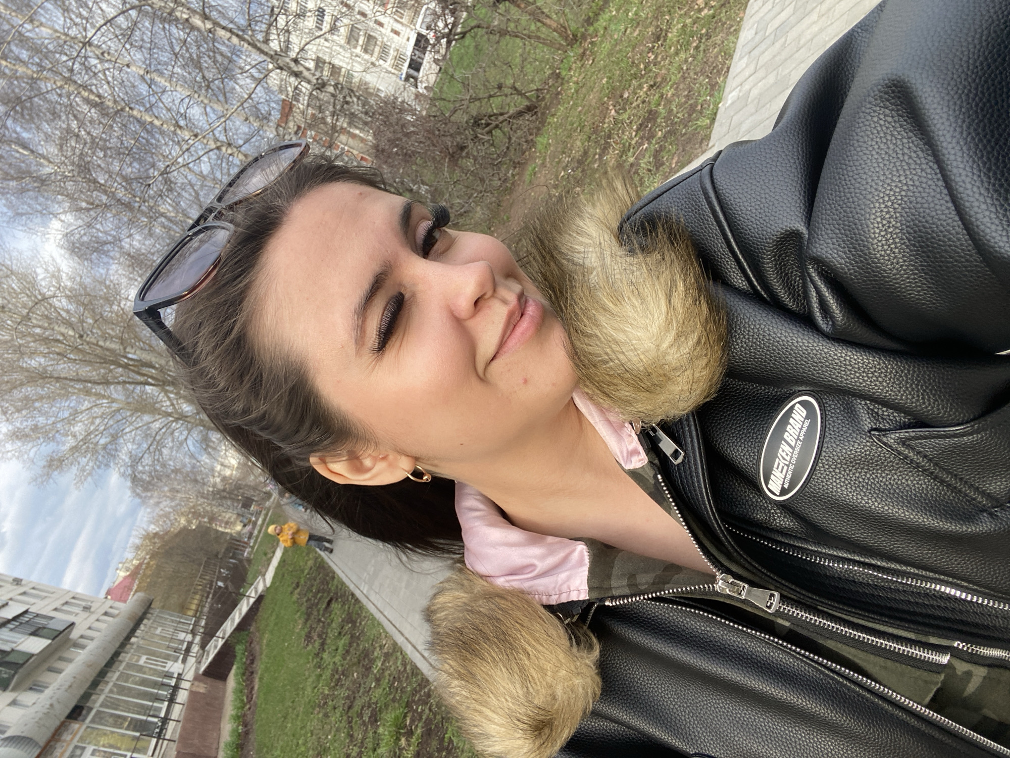 A young woman is depicted in a close-up selfie, taken outdoors in what appears to be Chaikovsky, Russia. She is shown from the chest up, looking towards the camera with a subtle smile. Her dark hair is pulled back, and a pair of sunglasses rests on her head. She has prominent eyelashes and wears a small hoop earring in her left ear.
She is dressed in a textured black faux leather jacket with a fluffy, light brown fur collar. Underneath the jacket, a light pink garment is visible, with a camouflage-patterned fabric peeking out beneath it. A black oval patch on her jacket clearly displays the text "MAIKEEN BRAND" and "AUTHENTIC OVERSIZE APPAREL".
The background suggests a late winter or early spring setting. Leafless trees with intricate branches fill the upper part of the frame against a bright, slightly overcast sky. Patches of green grass are visible on the ground, alongside a grey paved path. In the middle ground, parts of a multi-story residential building can be seen. Further down the path, a person wearing a bright yellow jacket is visible in the distance. The overall lighting indicates daytime. The scene captures a casual, calm moment outdoors.