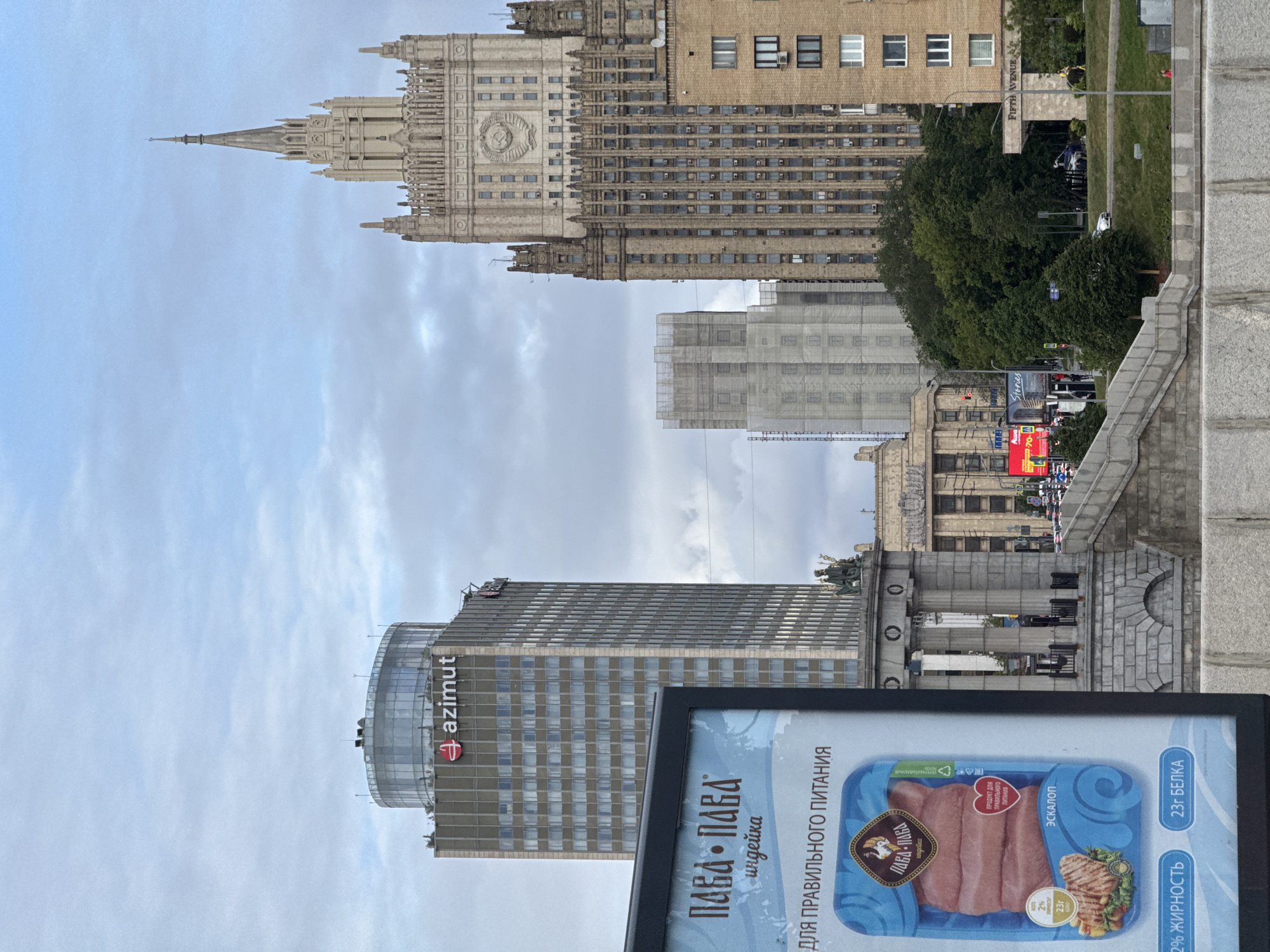 The image shows a partial view of the cityscape of Moscow, Russia, under a partly cloudy sky. In the background, the Ministry of Foreign Affairs building, one of the Seven Sisters skyscrapers, is prominent, with its tower extending into the sky. In the mid-ground, there is a modern high-rise building bearing the "azimut" logo. A large advertisement in the foreground features a product image of packaged turkey escalope. The advertisement's text is in Russian. Visible street signage includes "FIFTH AVENUE". Pedestrians and vehicles are seen on the street. The photograph appears to be taken from an elevated position, possibly a pedestrian bridge or elevated area.