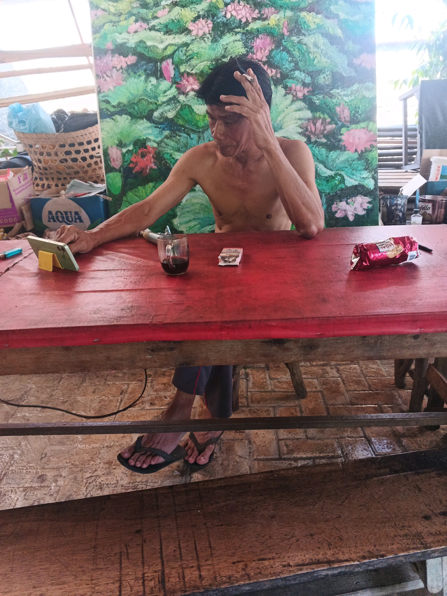 An individual, appearing to be an adult male with dark hair and a bare torso, sits at a red-painted wooden table. He holds a cigarette in his right hand, which is raised to his forehead, and appears to be looking downwards. In front of him on the table are a glass mug with a dark liquid, a mobile phone propped on a yellow stand, and a pack of cigarettes. A red, foil-wrapped package, possibly food or cigarettes, is also on the table to his right, along with a pen. To the left of the table, a large painting featuring pink and green lotus flowers is visible. Behind the man, a wicker basket contains light blue plastic bags and other items. The scene is set outdoors, with a tiled floor visible beneath the table and what appears to be wooden benches. The ambient lighting suggests it is daytime. The location is identified as Ungaran, Indonesia.