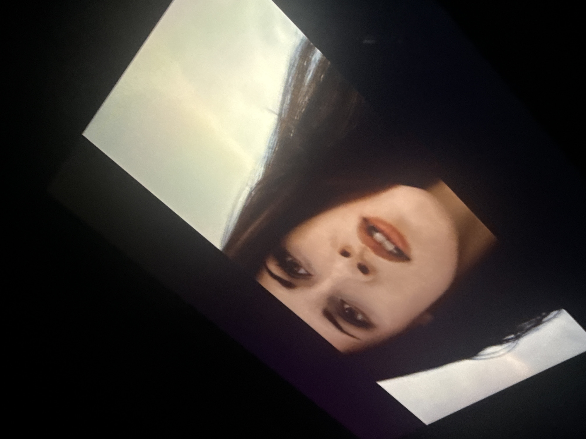 The image shows a section of a screen, likely a monitor or television, displaying a woman's face. She has fair skin, dark hair, and wears red lipstick. The background behind her appears to be a pale sky. The screen is at an angle in a dark setting. The location is not determinable from the image.