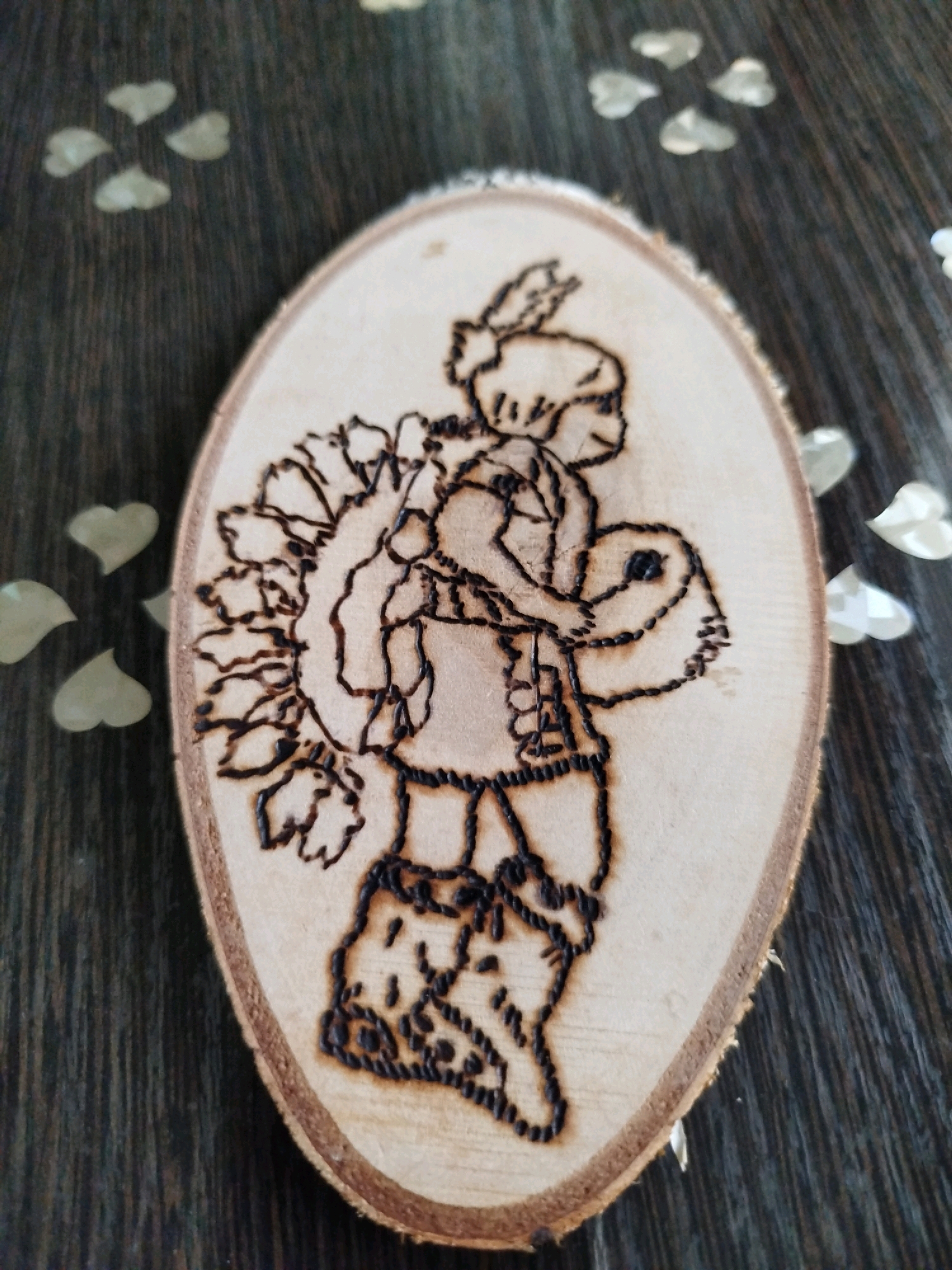 A pyrographic portrait of a character, possibly a Native American, is etched onto an oval piece of wood. The character is depicted from the chest up, facing left, with a headdress of feathers. They are wearing a fringed garment and high boots. The background is a dark, textured surface, possibly wood or fabric, with scattered heart-shaped confetti in a light green hue. The image is taken from a slightly elevated angle, with the oval wood piece tilted. No specific location in Крывасельскі сельскі Савет, Belarus is identifiable from the image itself.