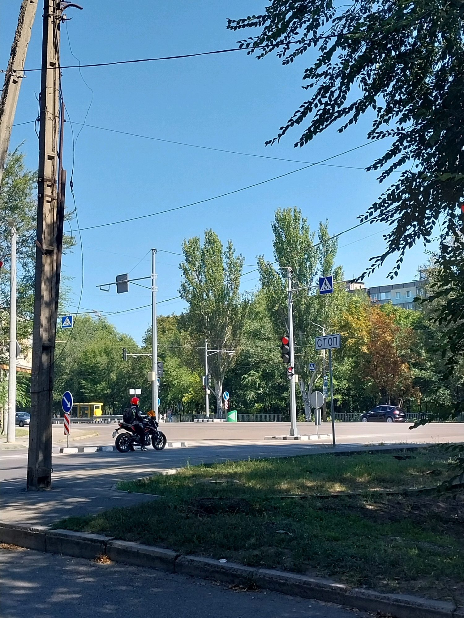 This is a medium-angle, eye-level, full outdoor shot of a street intersection in Dnipro, Ukraine, on a clear, sunny day. In the foreground, a concrete sidewalk runs along the bottom of the frame, with grass and some patches of dark soil on the right. A paved road occupies the middle ground. A motorcycle with a rider wearing a red helmet is stopped at the intersection. A traffic light with red illumination is visible on a pole to the left of the motorcycle. A "STOP" sign is mounted on a pole to the right of the traffic light. Further back, a yellow bus is parked behind a fence. Various trees line the background, some with green leaves and others with autumnal yellow and orange foliage. A multi-story building is visible behind the trees on the right side of the frame. Power lines and poles are prominent features throughout the image.